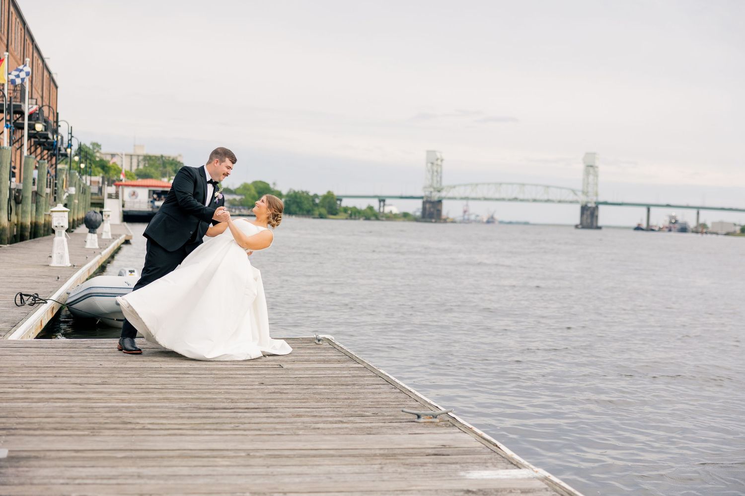 Groom dips bride in white gown on waterfront dock with bridge in background.