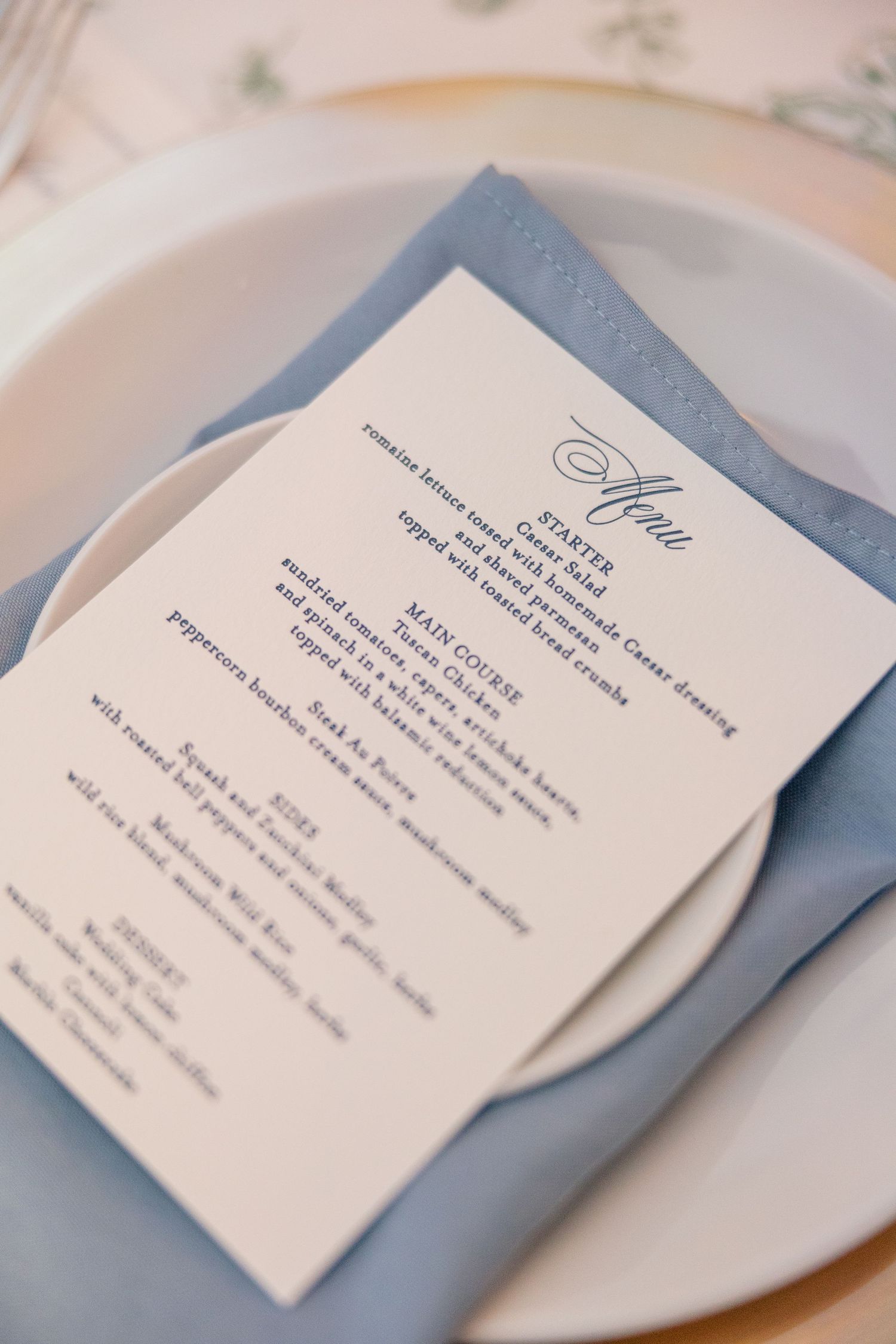 Elegant wedding dinner menu card rests on blue linen napkin atop white plate setting.