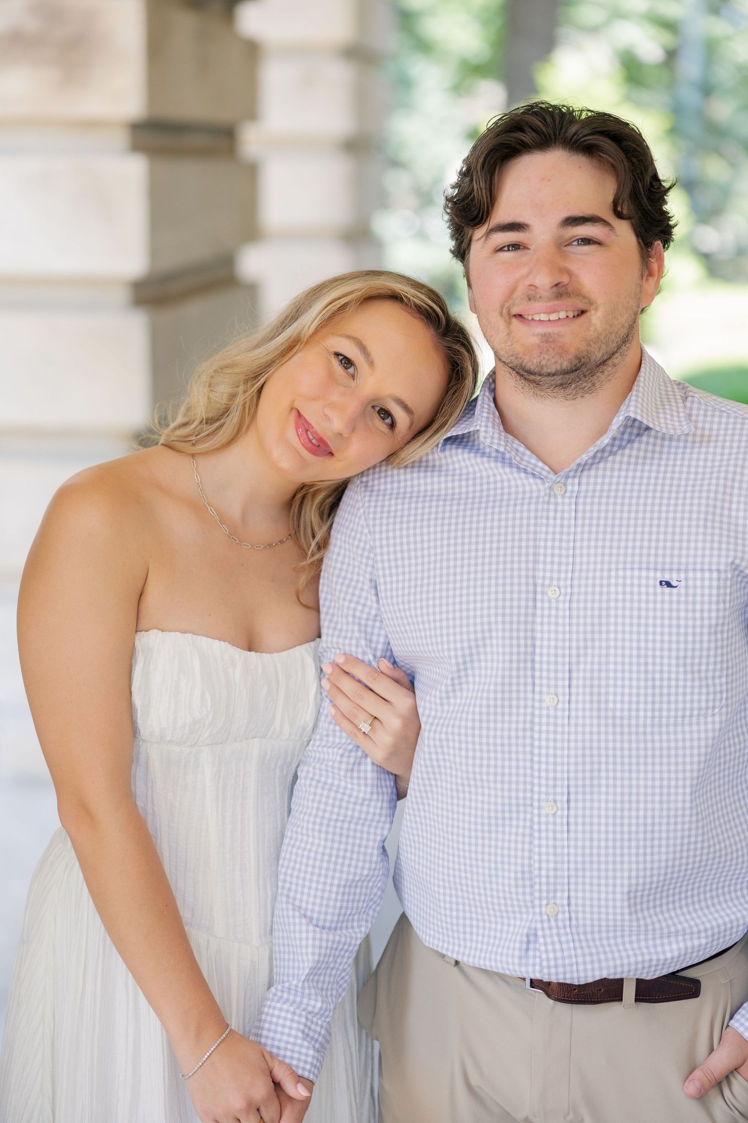 Smiling couple posing together, woman in white strapless dress resting head on mans shoulder outdoors.