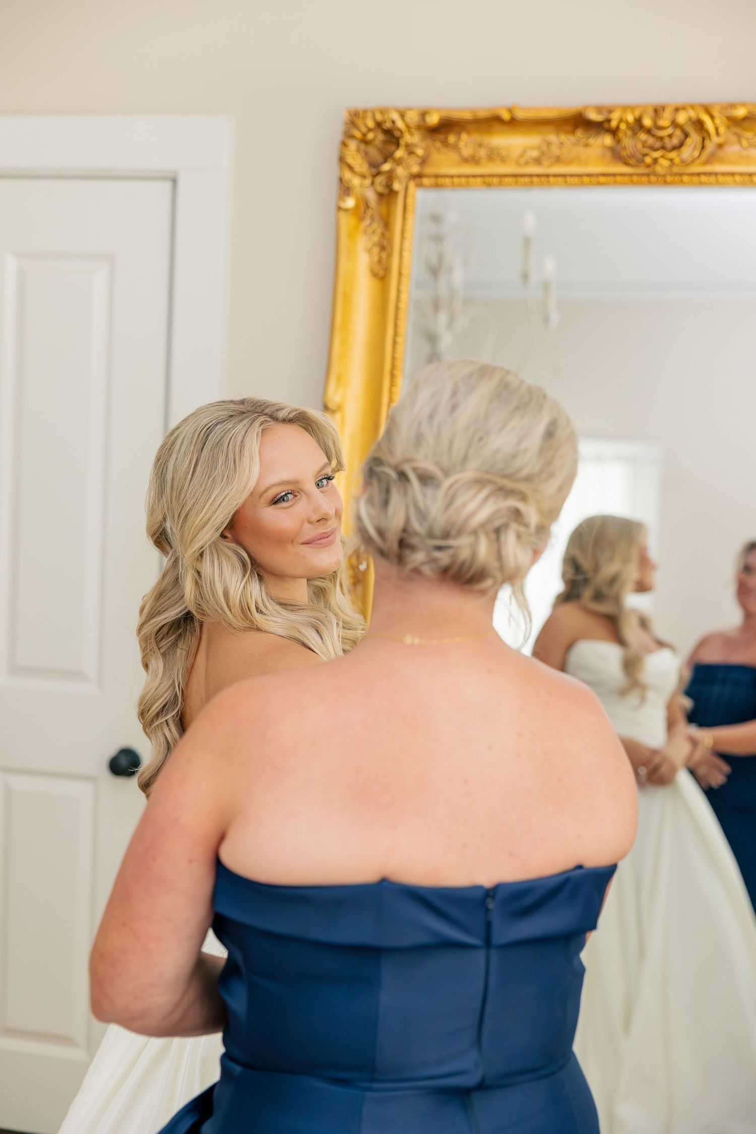 Bride smiling at older woman in blue strapless gown during wedding preparations in front of gold mirror.