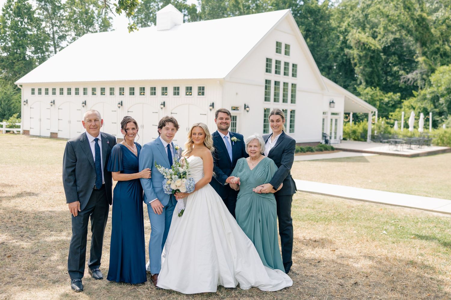 Wedding party poses in front of white barn venue with bride in ball gown and guests in blue attire.