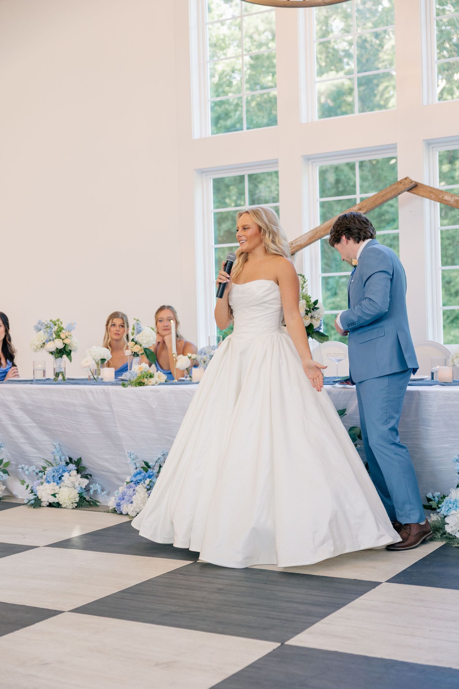 Bride in strapless ball gown holds microphone while groom in blue suit bows on checkered dance floor.