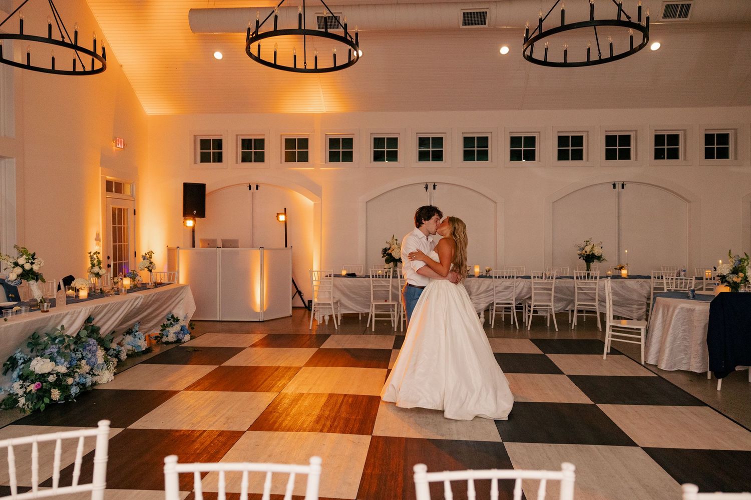 Bride and groom share their first dance on a checkered floor in an elegantly lit reception hall.