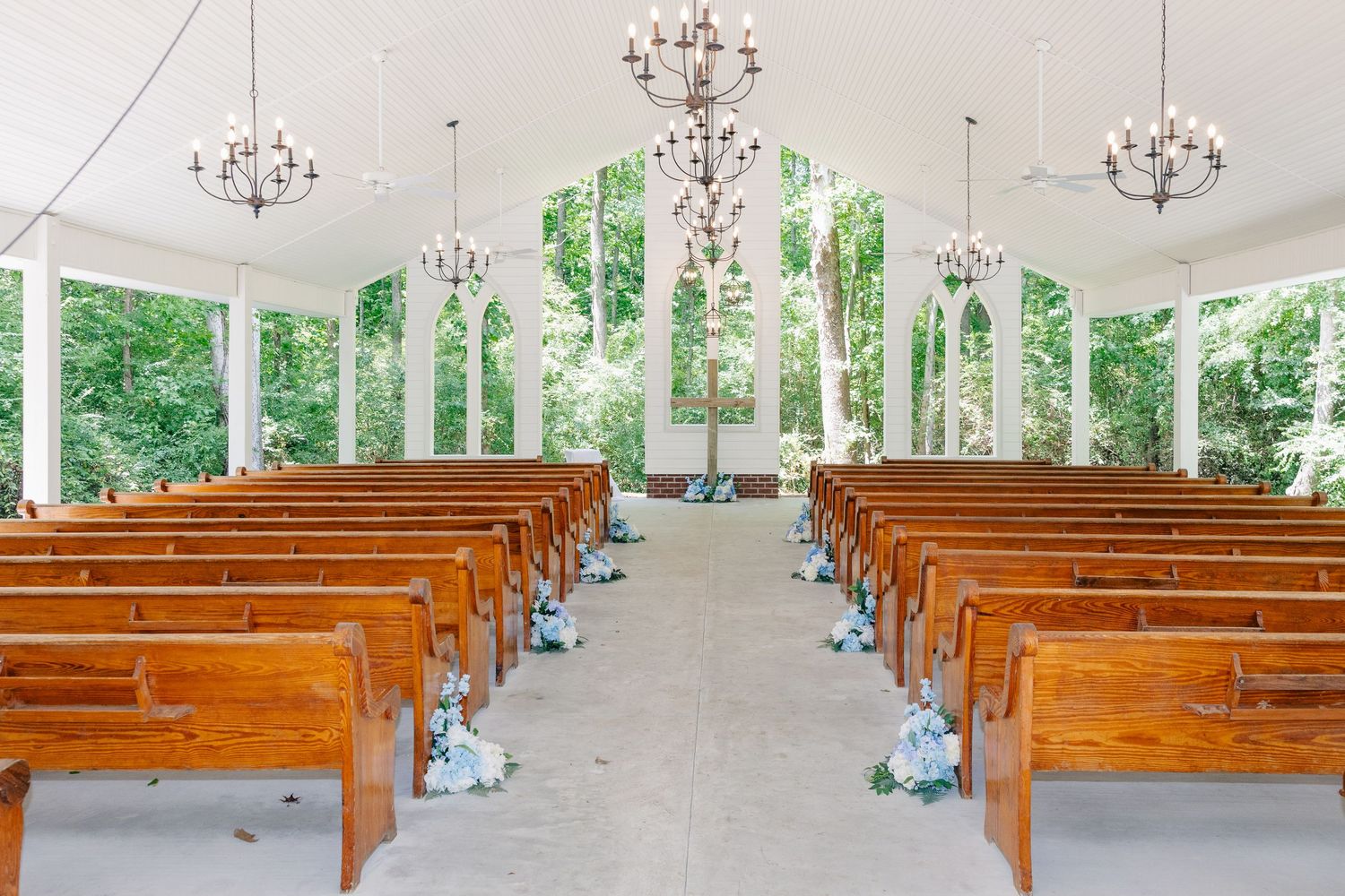 Elegant white chapel interior with wooden pews, chandeliers, and lush green views through windows.