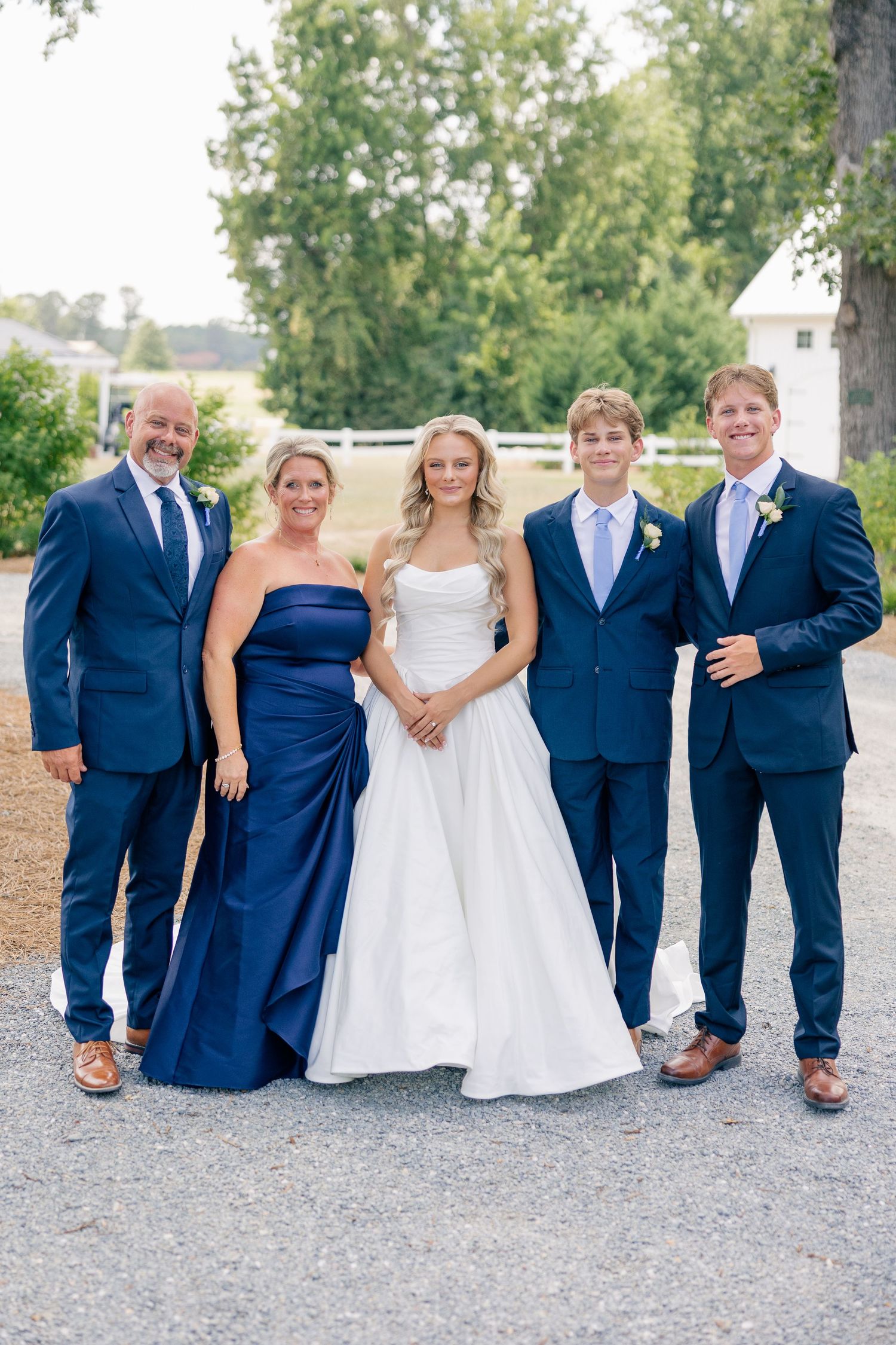 Bride in white gown poses with family in coordinating navy blue attire outdoors near white barn.