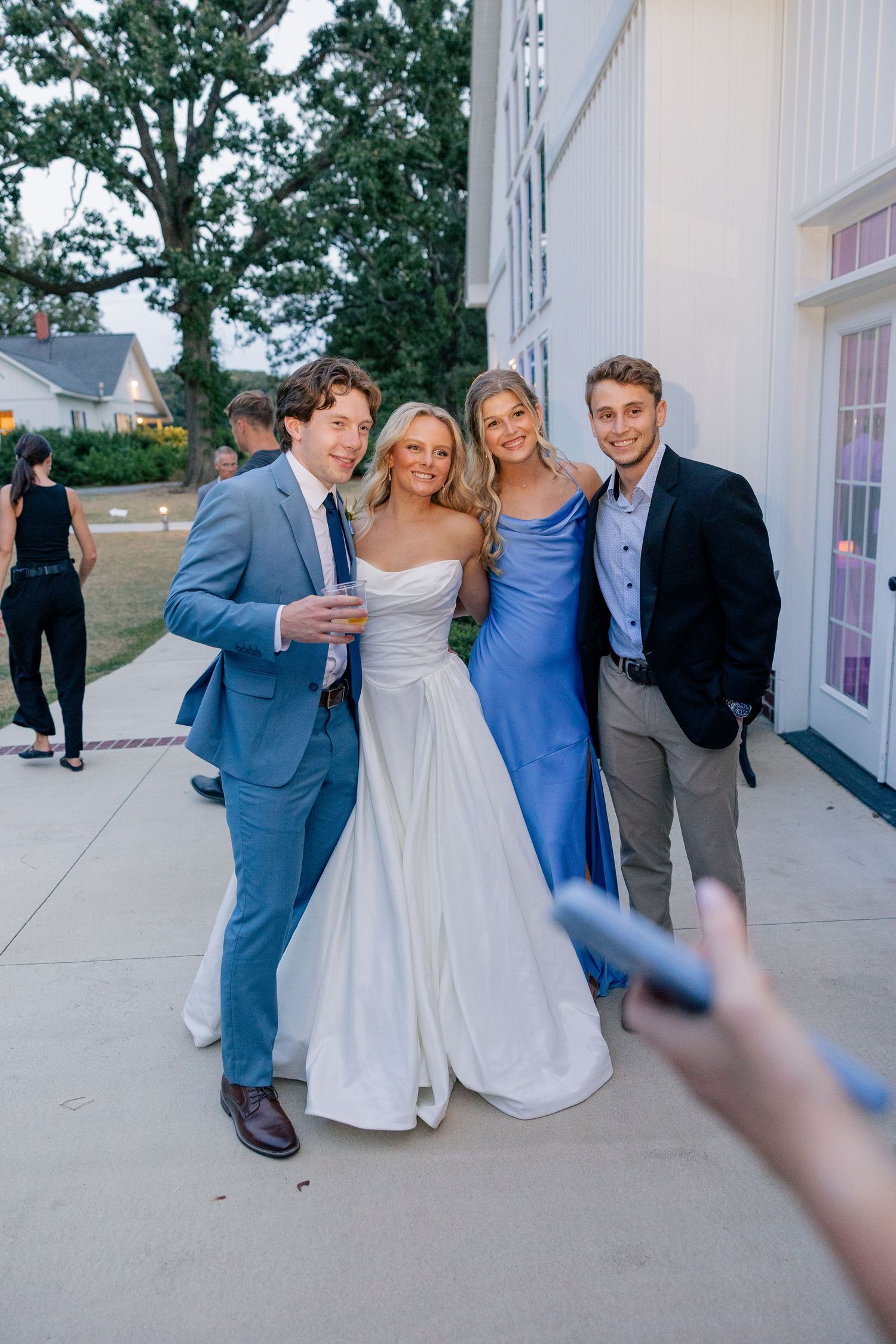 Bride in white gown and bridesmaid in blue dress pose with two men in suits outside wedding venue.