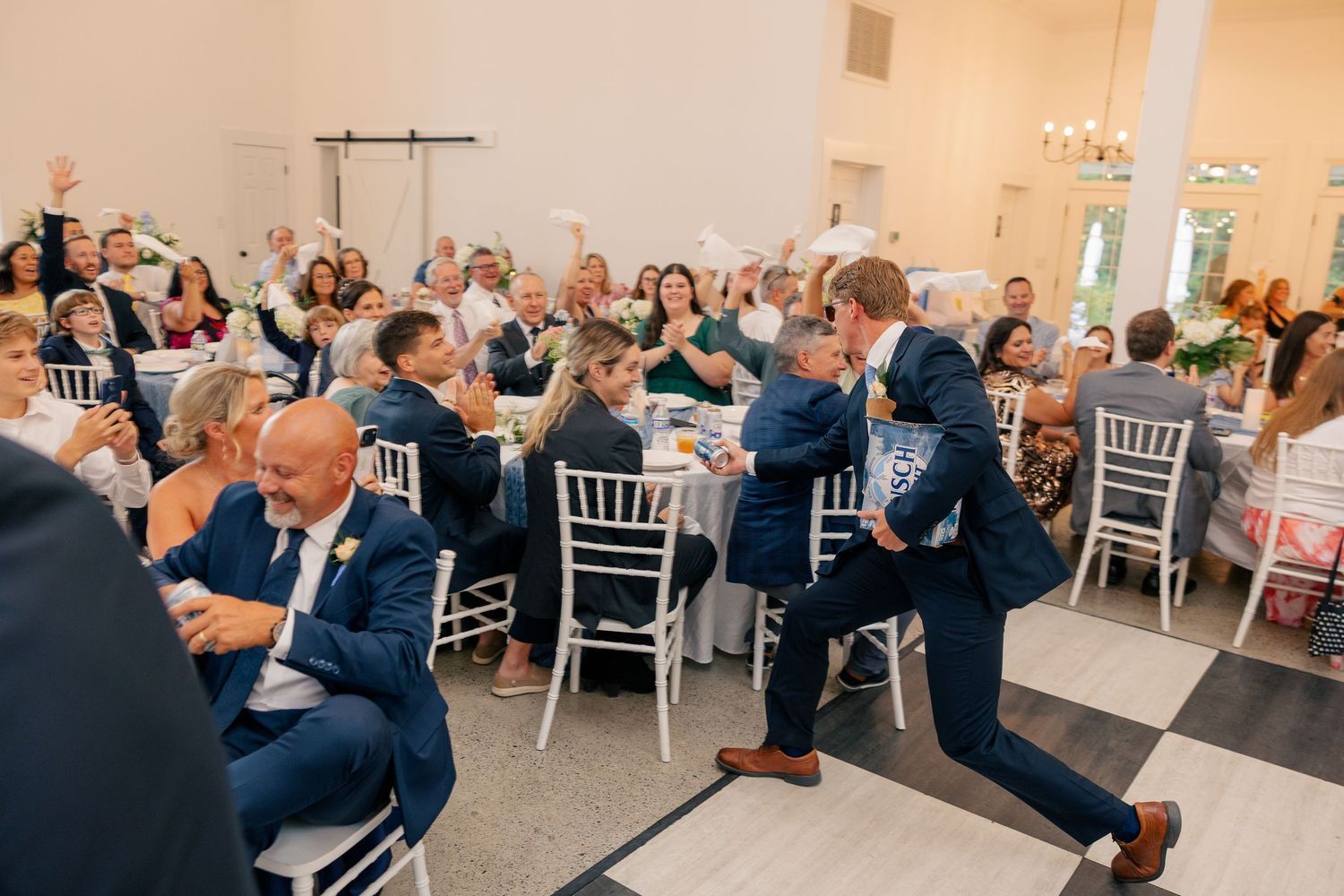 Groom dances energetically on checkered floor as wedding guests cheer during reception celebration.