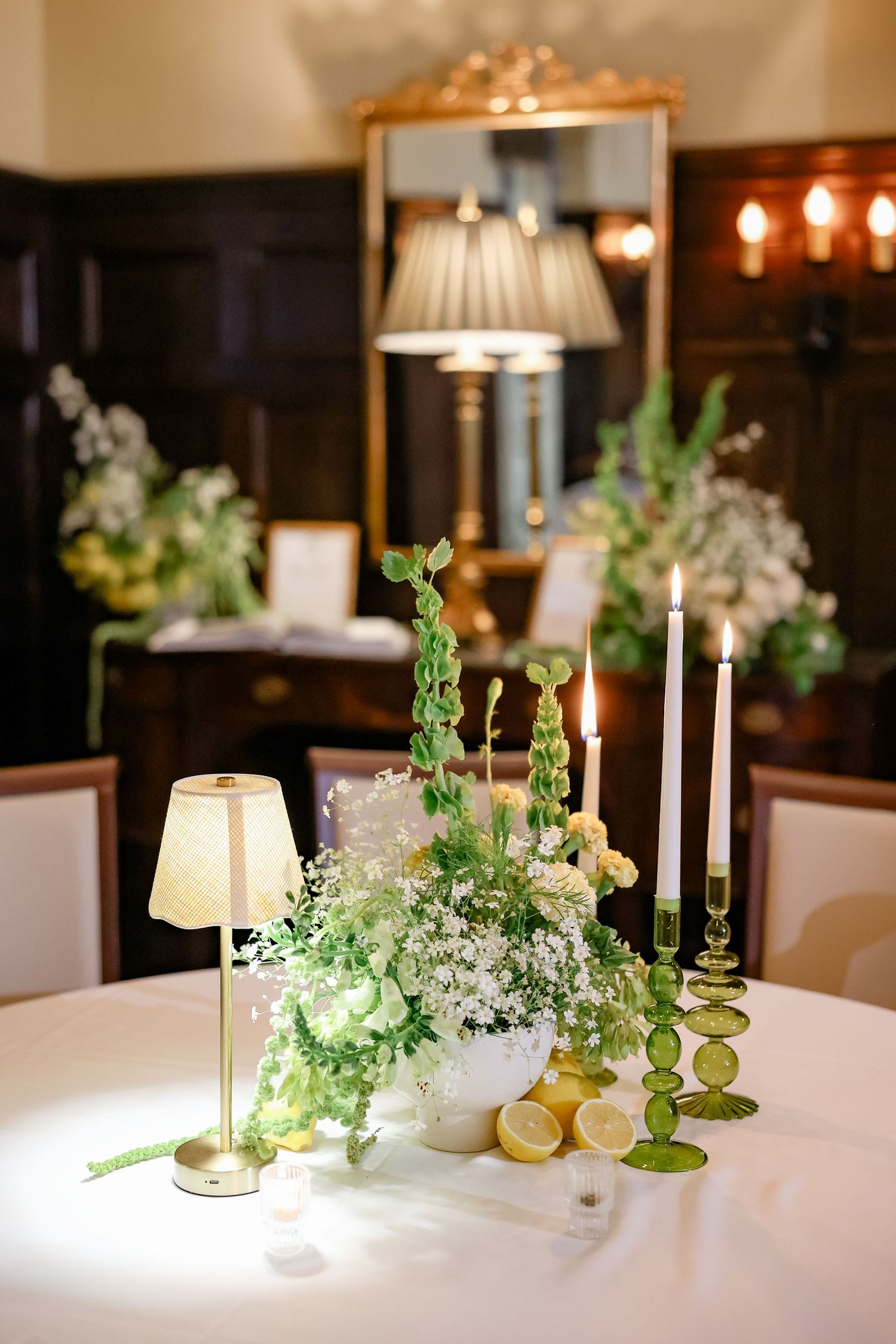 Elegant wedding reception table with green floral centerpiece, white candles, lemon accents, and a small lamp in a dark wood room.