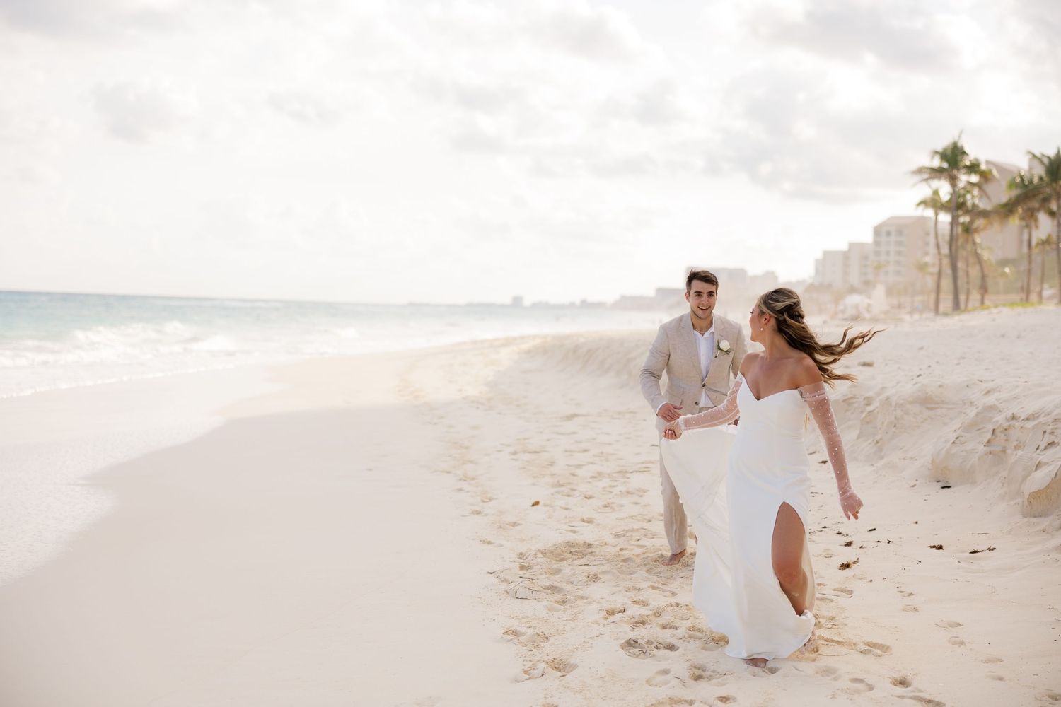Couple in white outfits walking and playing along a sunny beach shoreline with palm trees in the background.