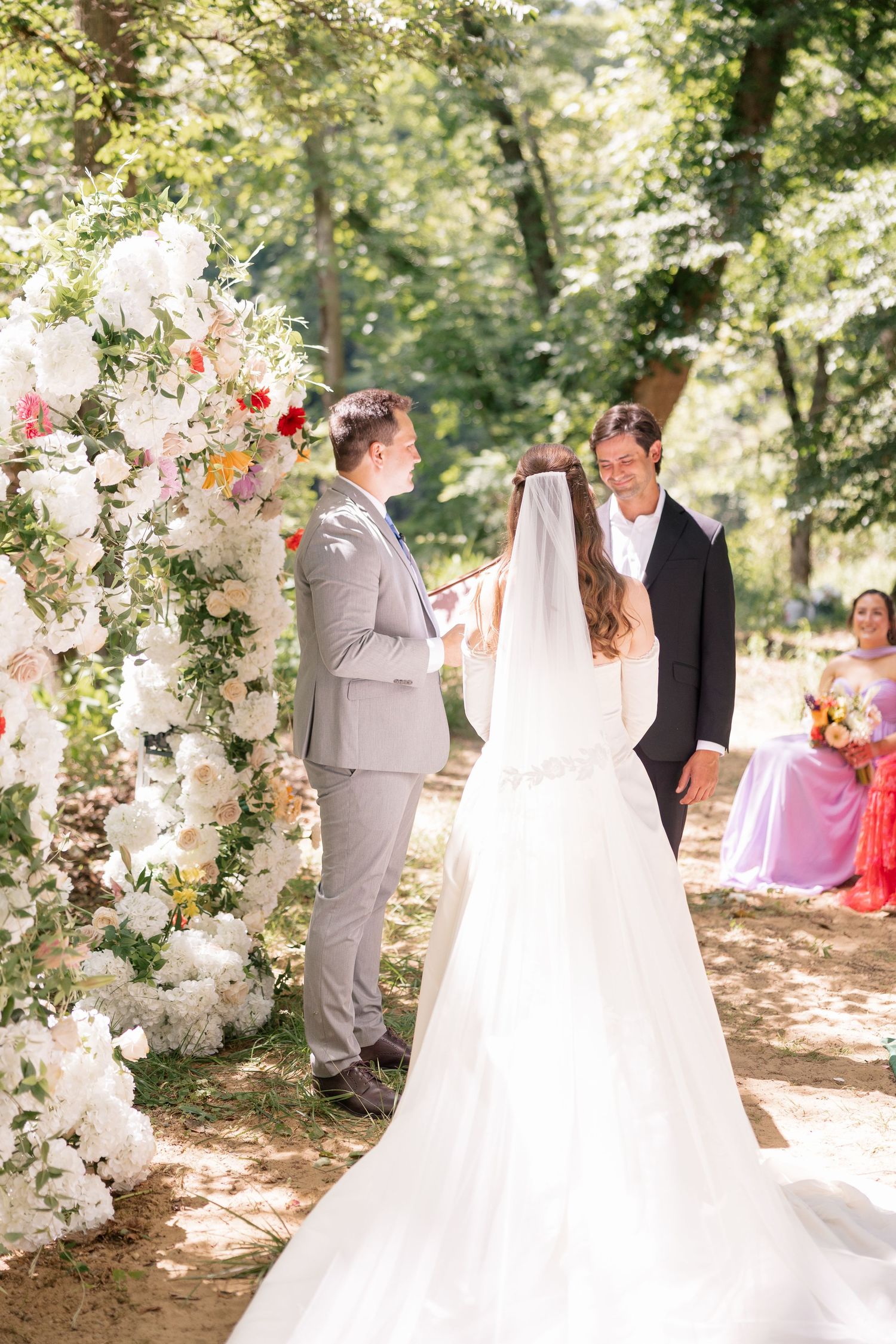 Bride and groom exchange vows at outdoor ceremony beside tall white floral arch in sunlit forest.