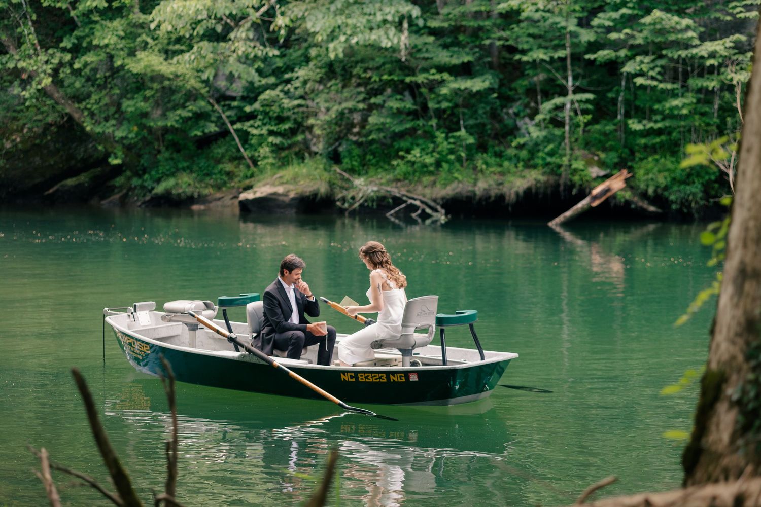 Bride and groom share a romantic moment in a rowboat on a serene green lake surrounded by lush forest.