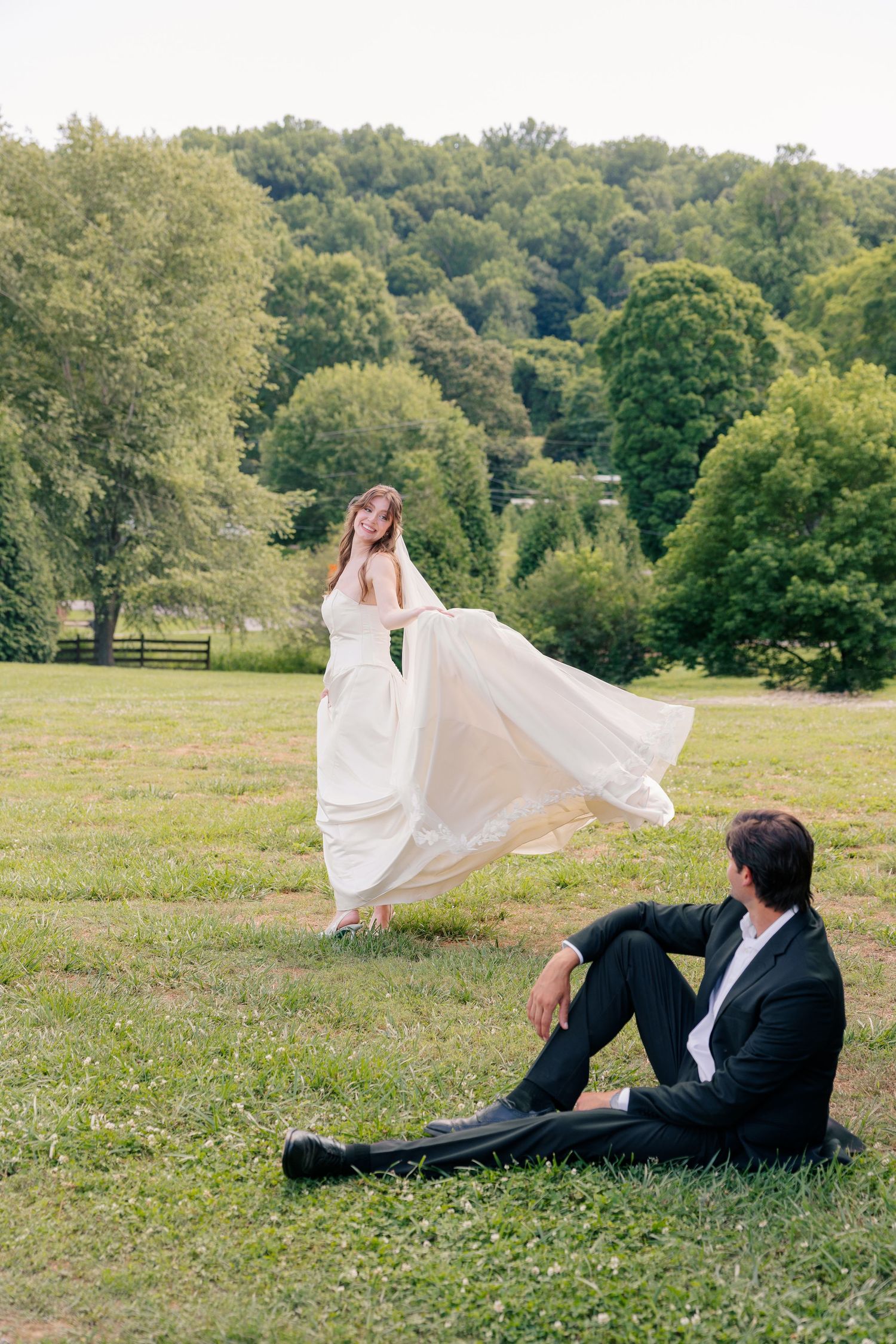 Bride twirls her flowing white gown in a green meadow as groom sits relaxed on the grass.