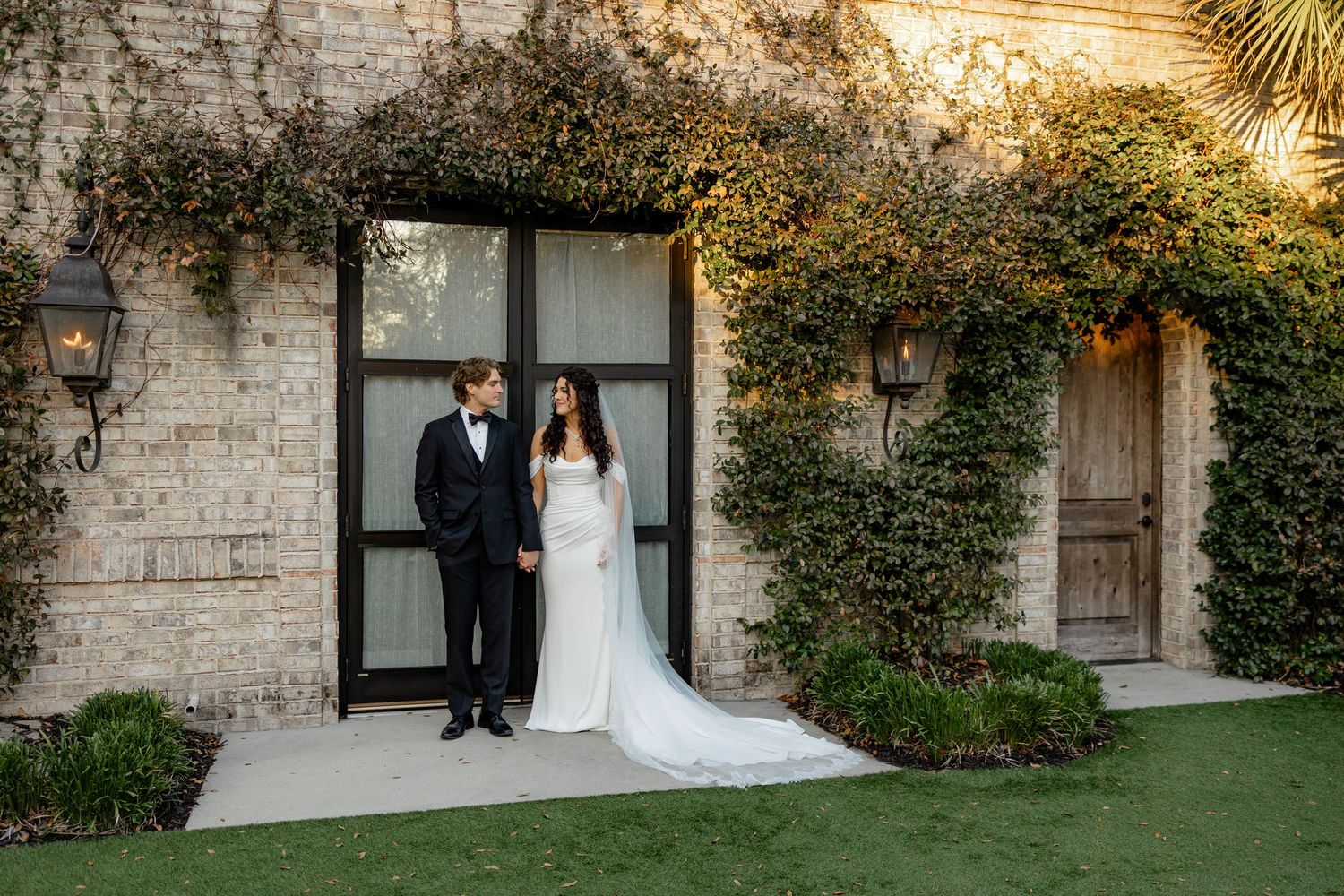 A couple stands in front of a stone building with climbing ivy and black framed windows during a sunset evening.