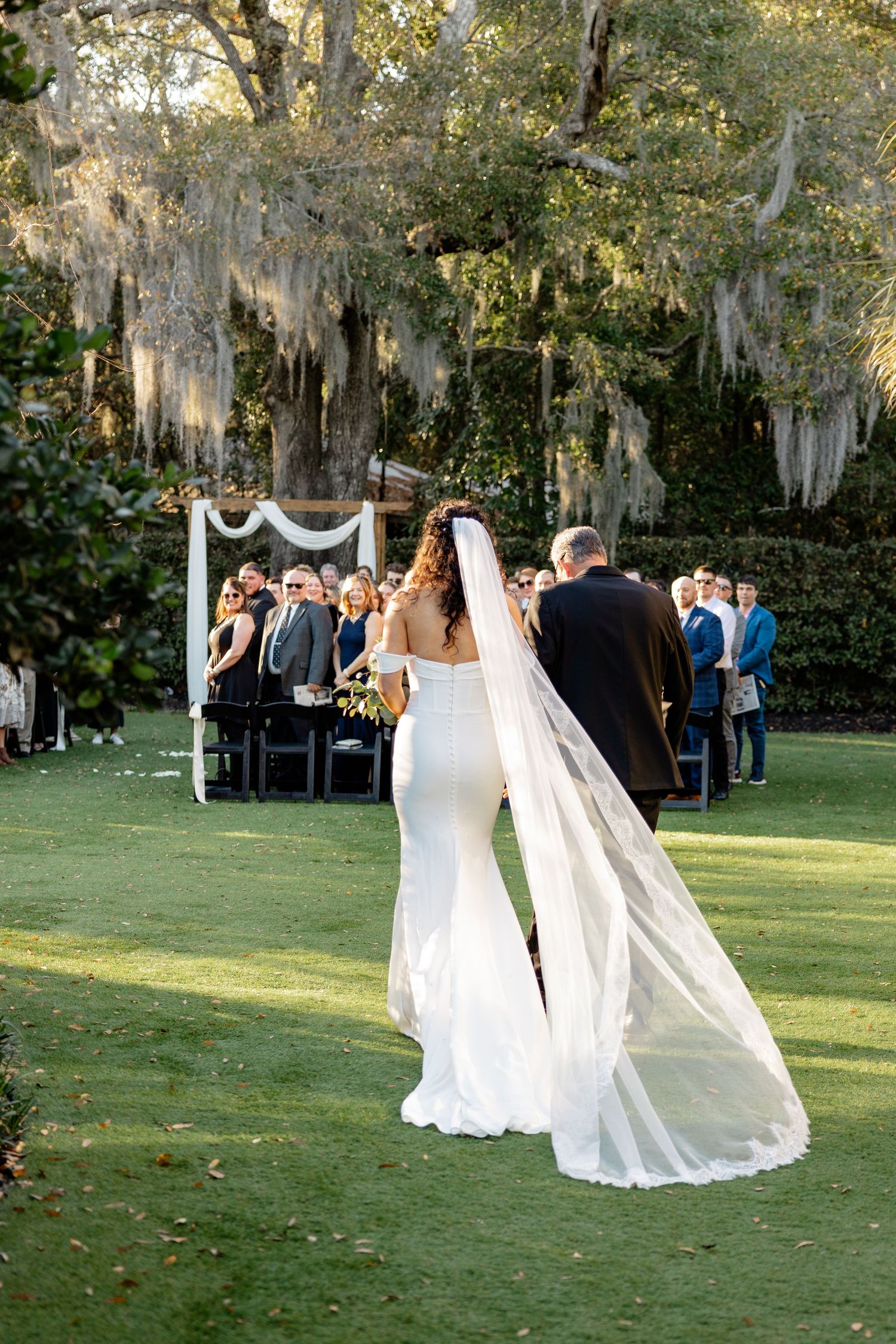 An outdoor wedding ceremony under Spanish moss draped trees with white fabric arch and guests seated on chairs.