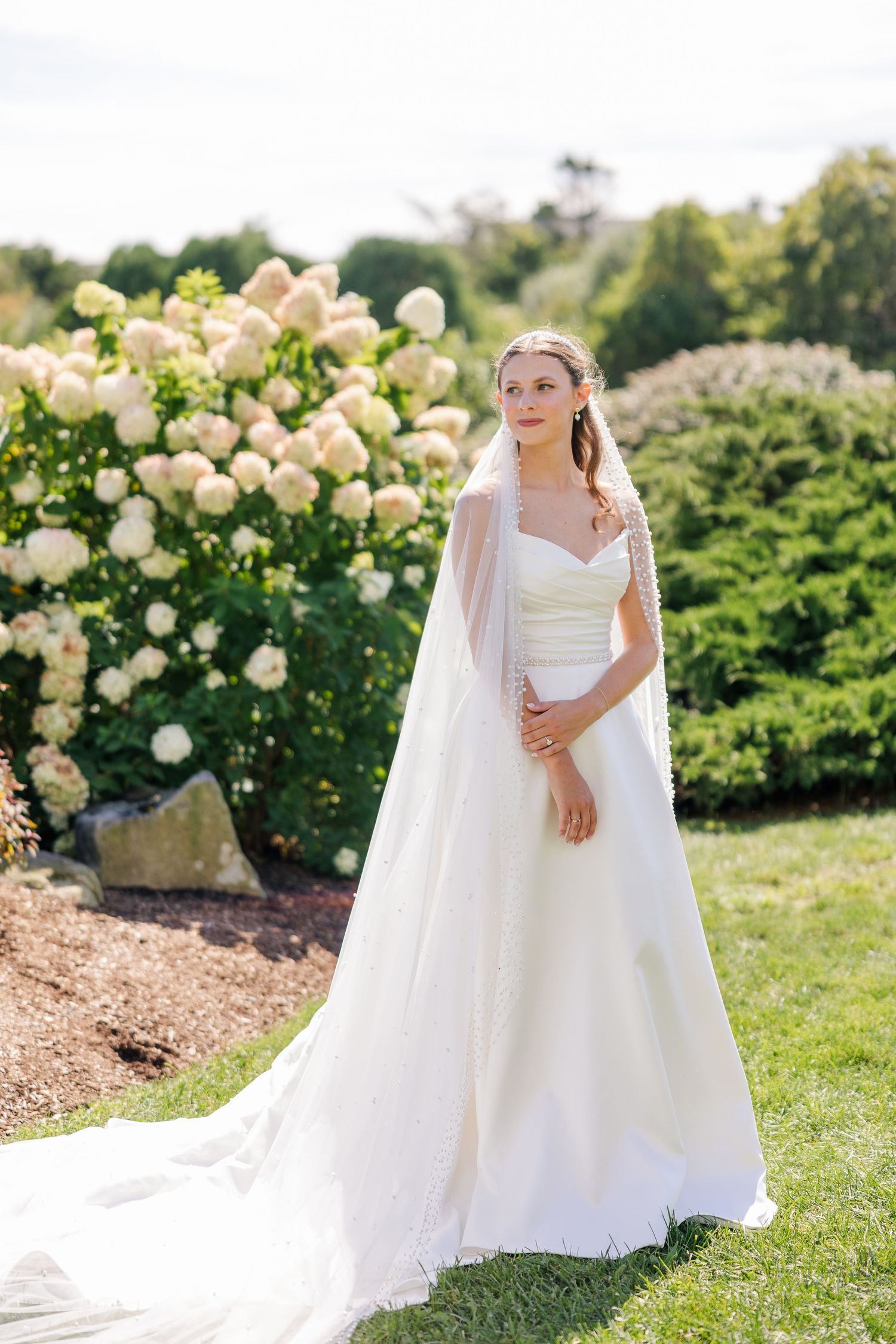 Bride in white ball gown with veil standing in garden with white hydrangeas.