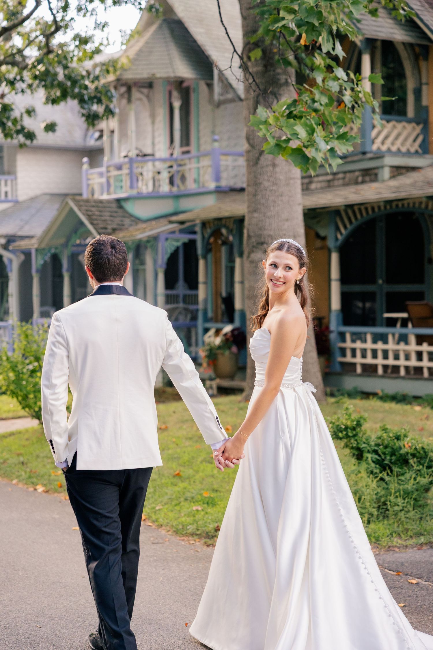 Bride in white gown looks back smiling while holding grooms hand walking past Victorian houses.