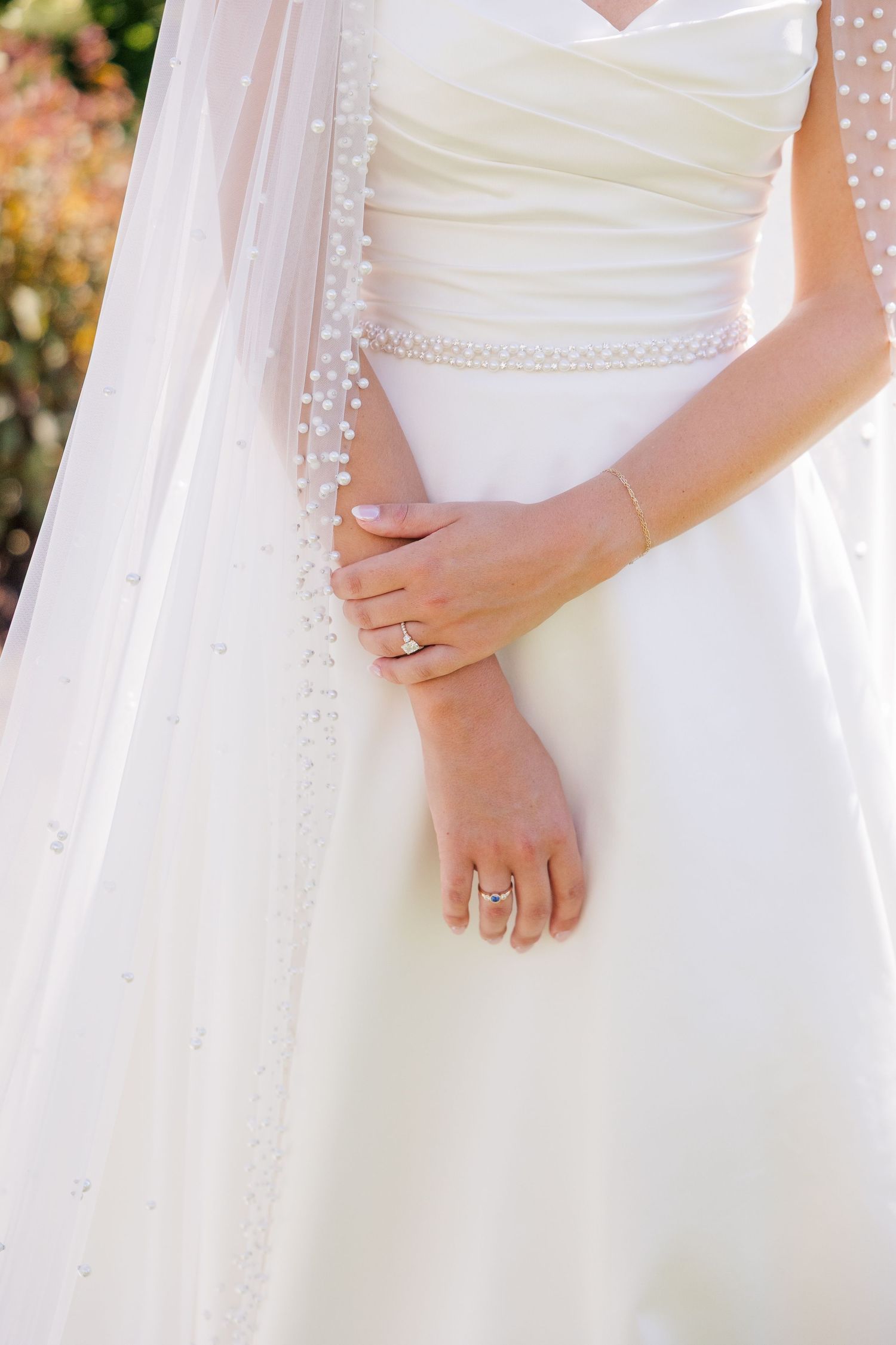 Bride in white wedding gown with pearl-trimmed veil, hands clasped showing elegant engagement and wedding rings.