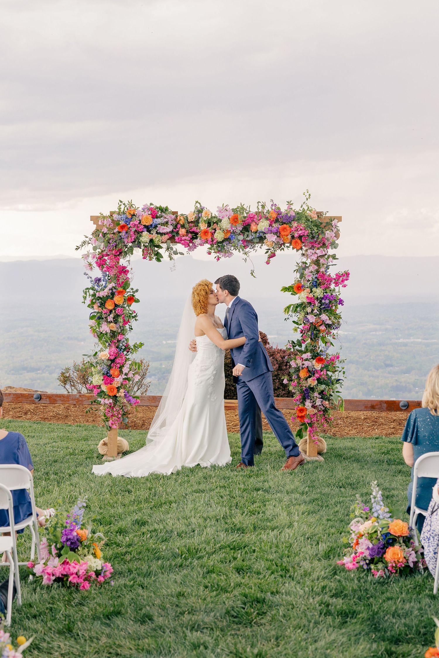 Bride and groom share first kiss under vibrant rainbow floral arch with mountain landscape backdrop.