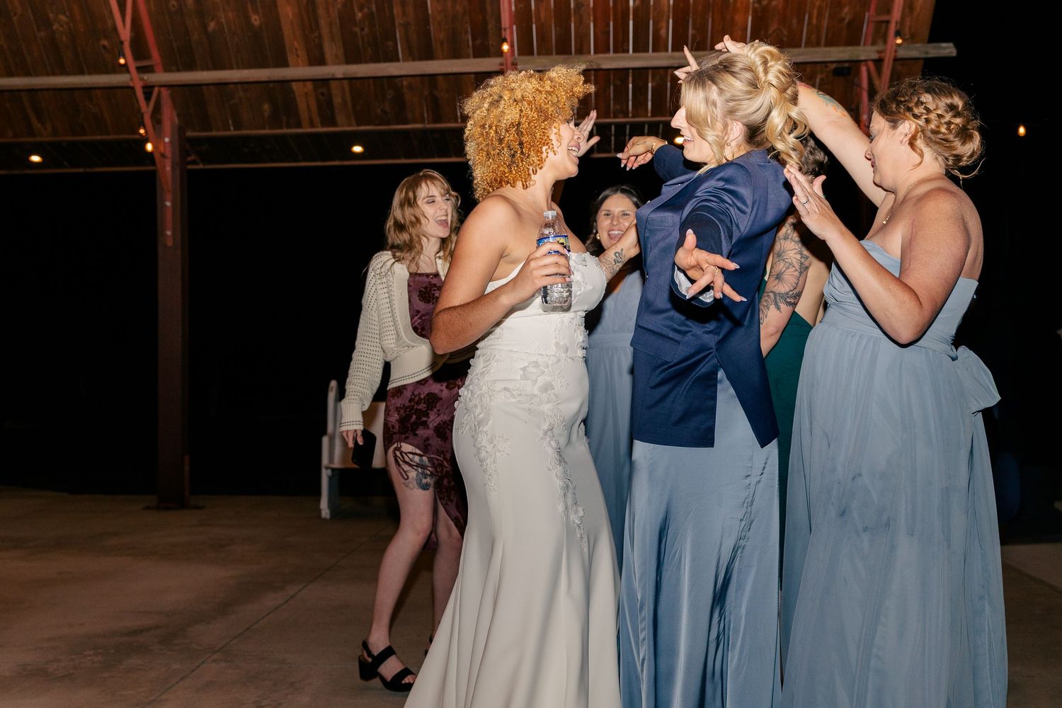 Bride in white gown dances joyfully with bridesmaids in blue dresses at lively indoor wedding reception.