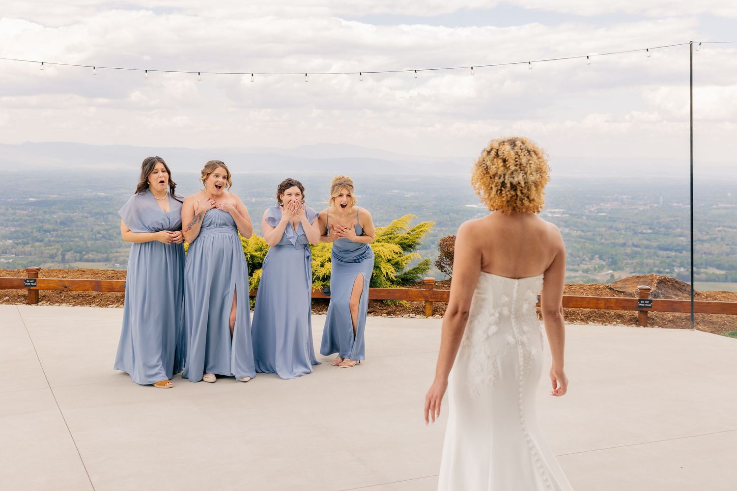 Bridesmaids in blue dresses react with surprise seeing bride in white wedding gown on terrace.