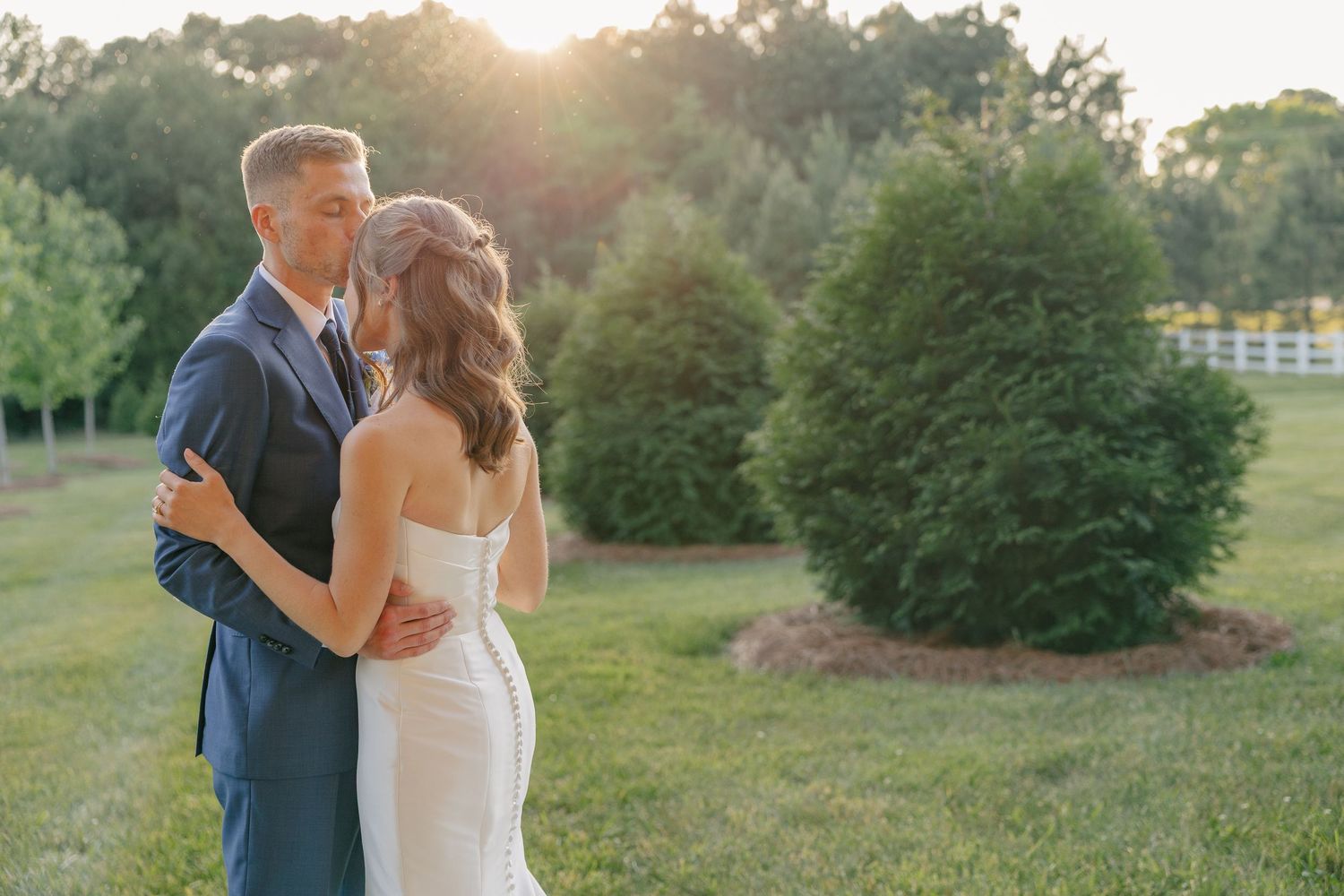 Bride and groom embrace during golden sunset in lush green outdoor wedding venue.