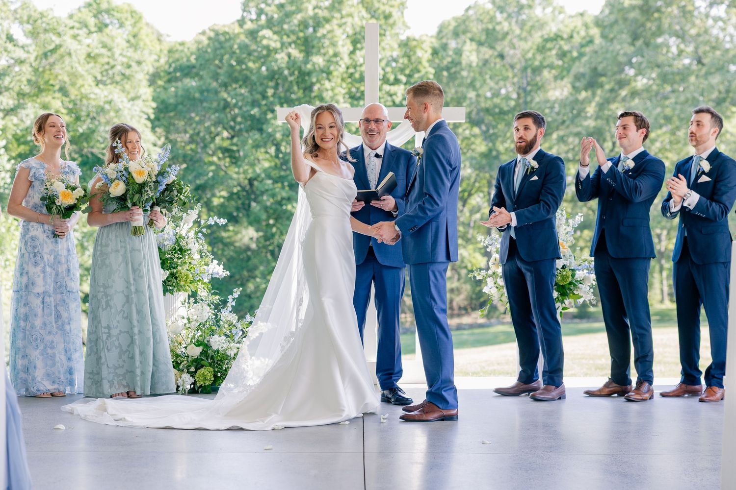Bride and groom celebrate at outdoor wedding ceremony with bridal party in navy suits and sage bridesmaid dresses.