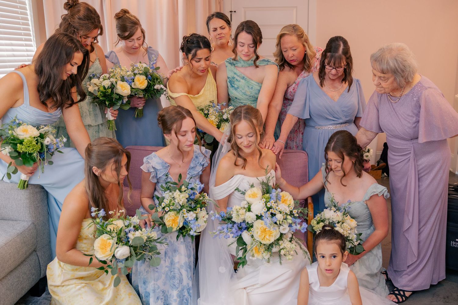 Bride surrounded by bridesmaids in blue, yellow, and lavender dresses holding spring bouquets indoors.