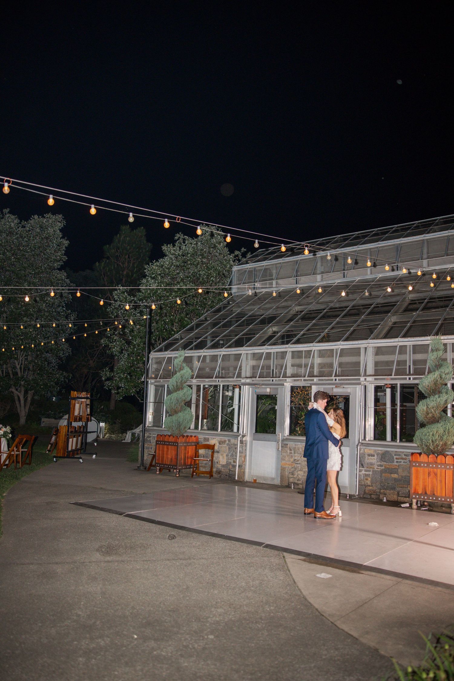 A couple shares an intimate moment outside a greenhouse under string lights at night.