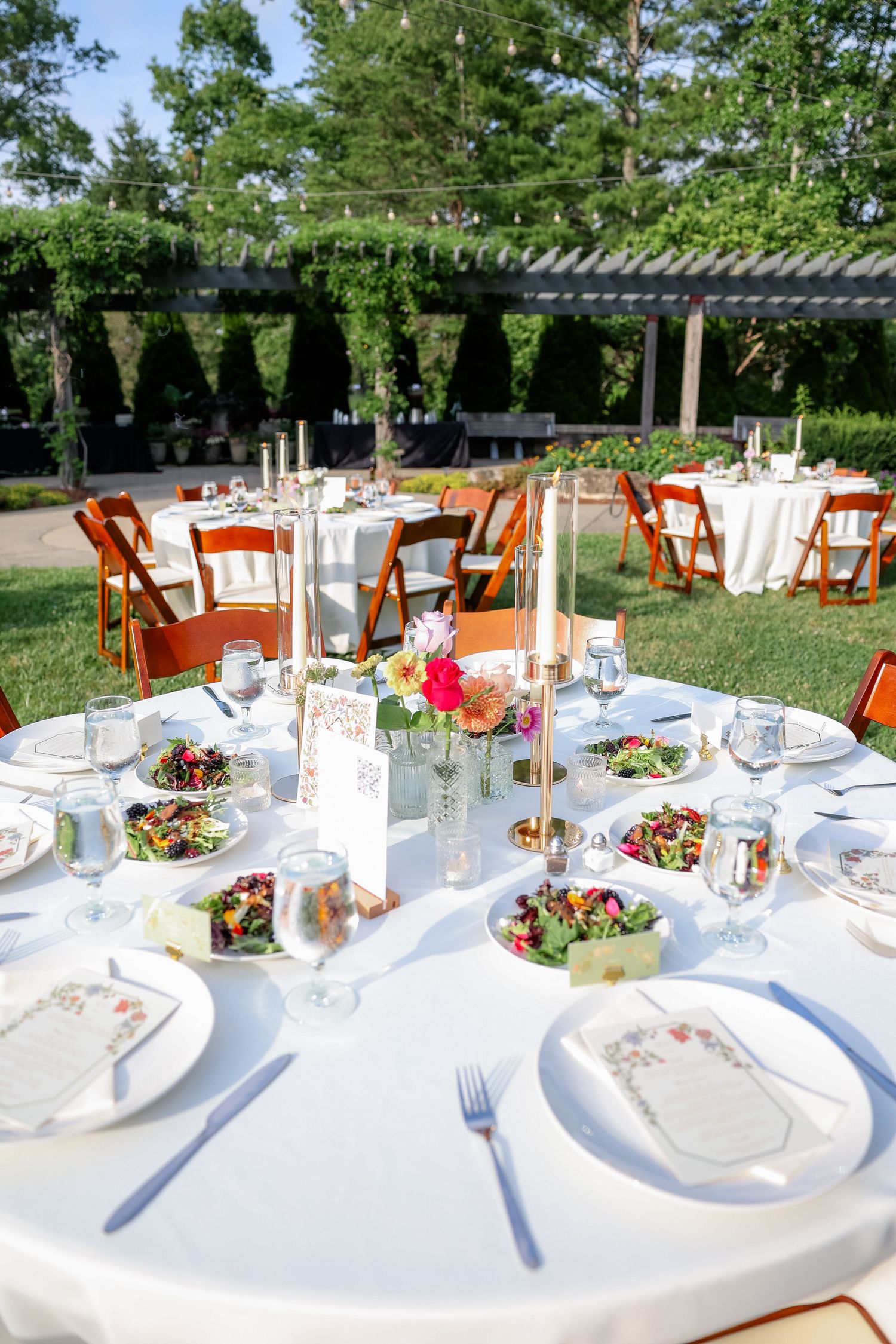 Outdoor wedding reception setup with round tables, white linens and wooden chairs under trees.