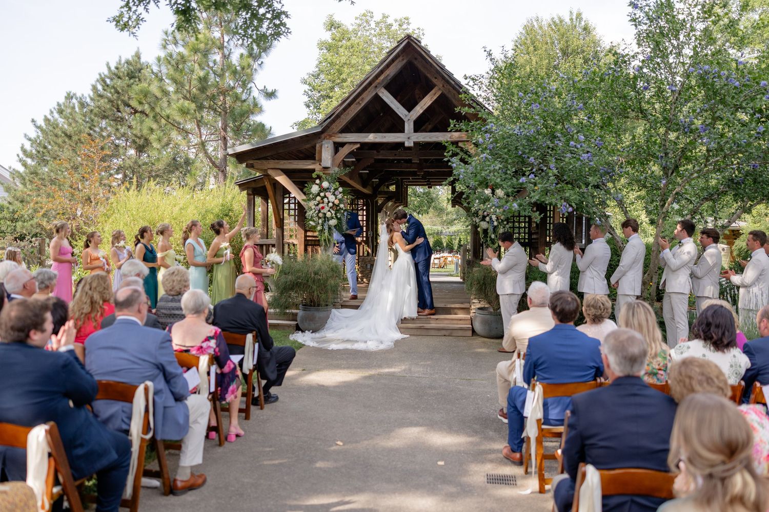 Outdoor wedding ceremony under rustic wooden pavilion with guests seated on white chairs.