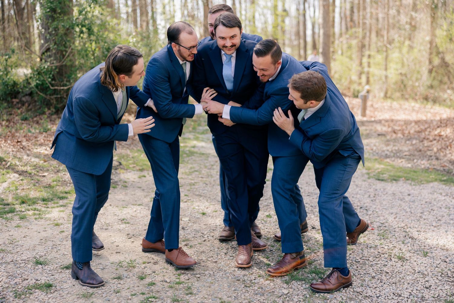 Groomsmen in navy suits pose playfully together on a wooded path for wedding photos.