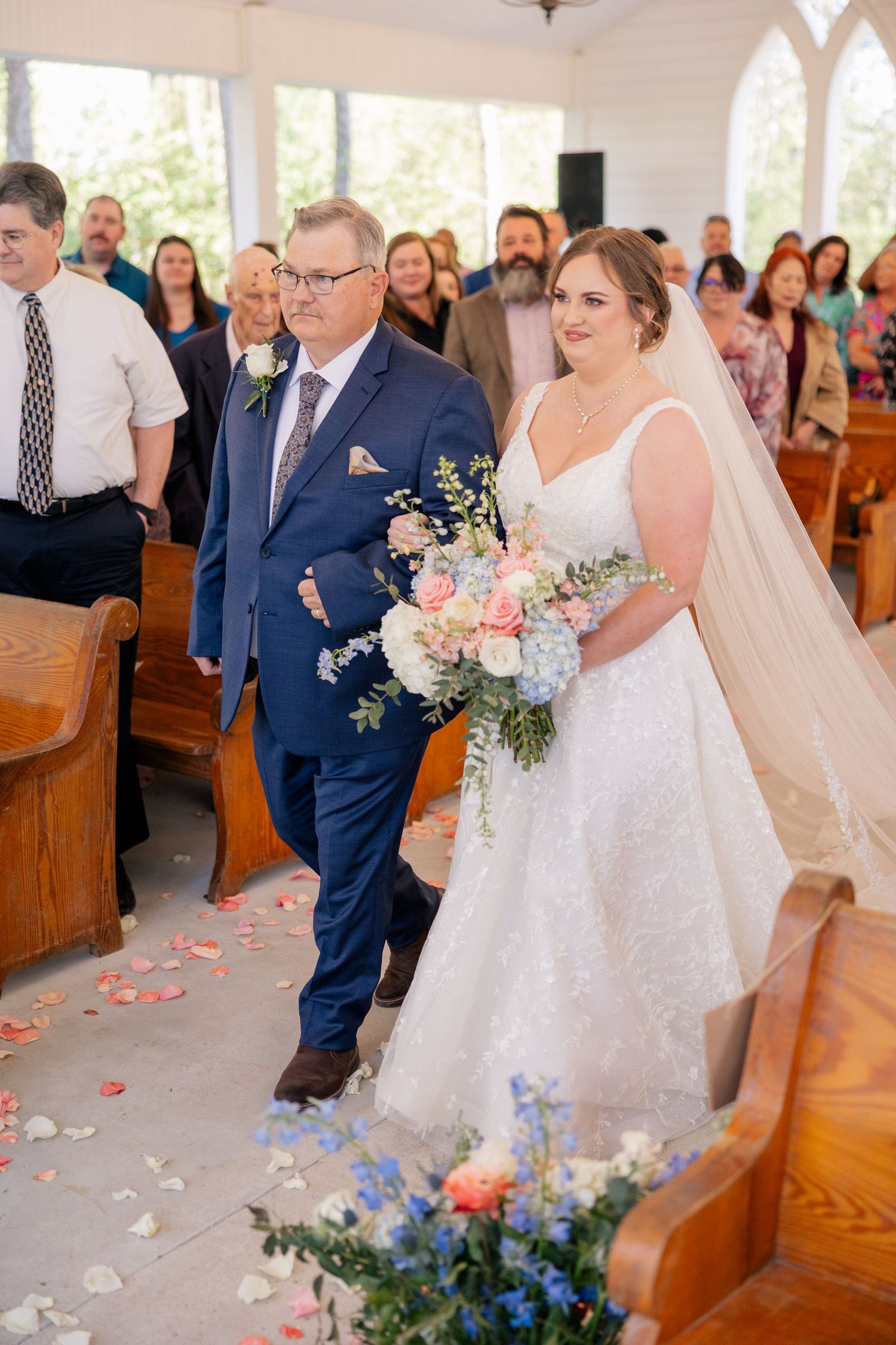 Wedding party processes down the aisle in a white chapel decorated with floral arrangements.