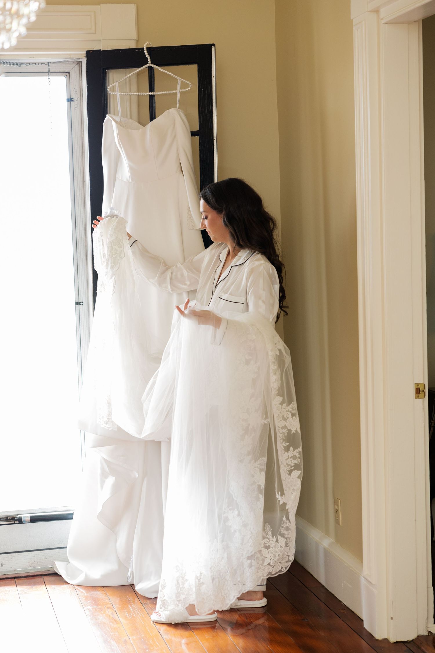 White bridal gown hanging in a sunlit room with elegant French doors and crystal chandeliers.