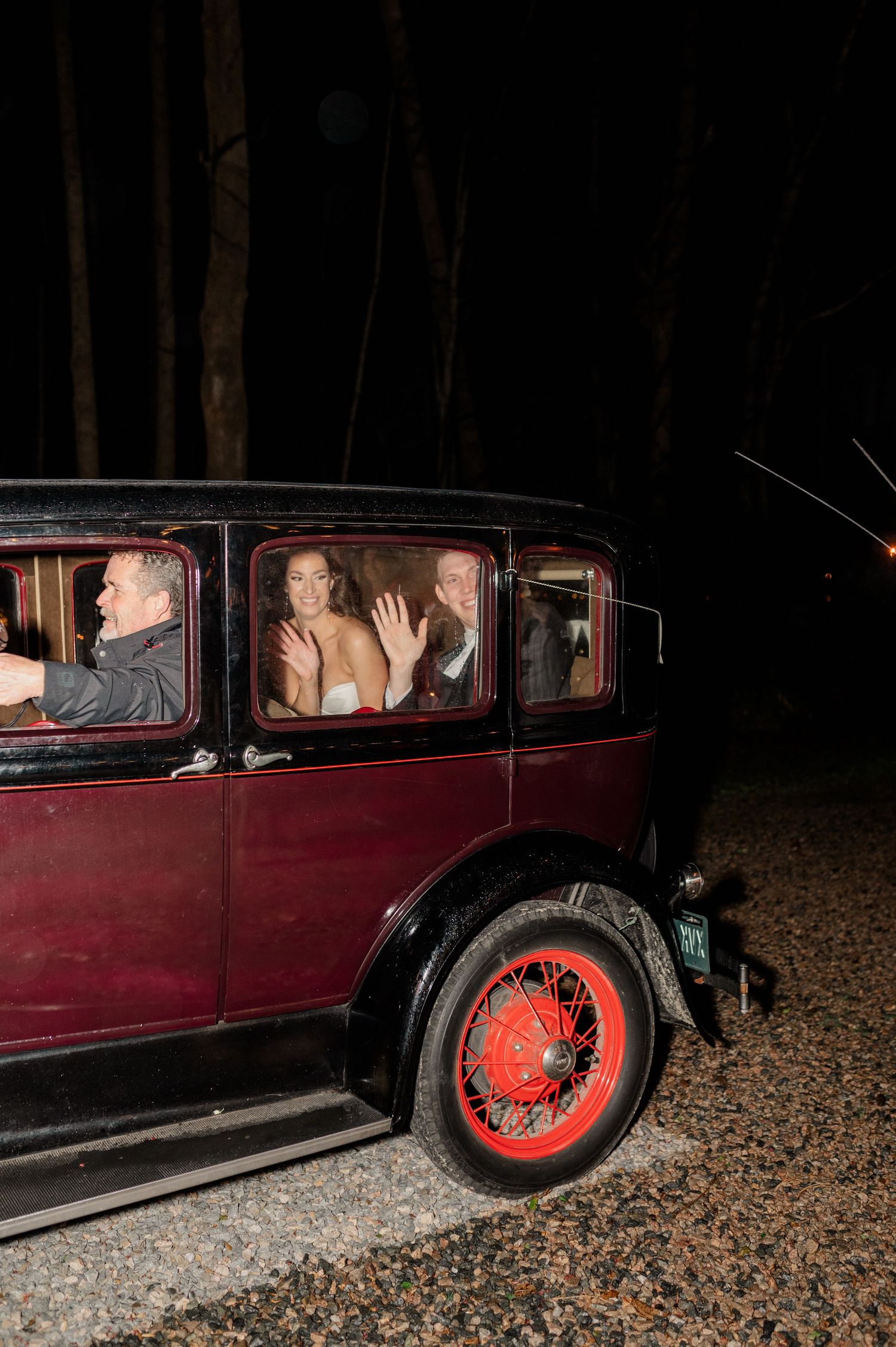 Vintage burgundy car with red wheels seen at night carrying wedding passengers.