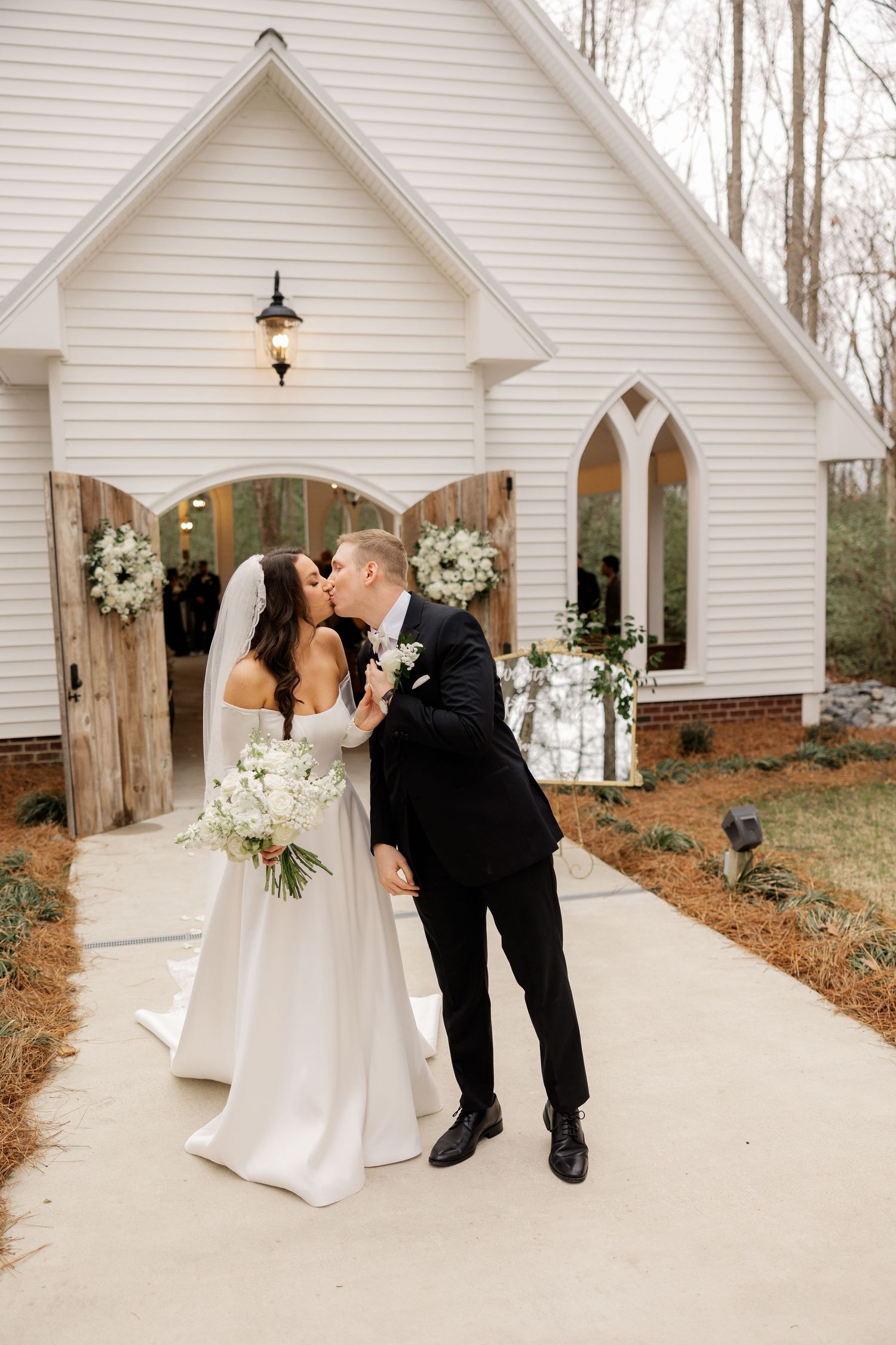 Romantic wedding moment outside charming white chapel with rustic wooden doors and floral decor.
