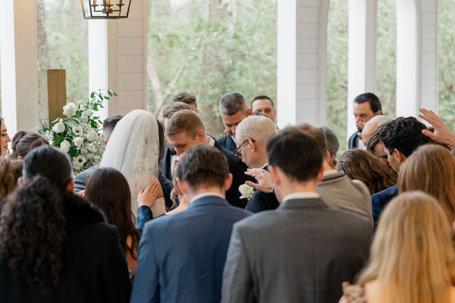Wedding ceremony guests gather in an elegant white chapel with tall windows and natural light.