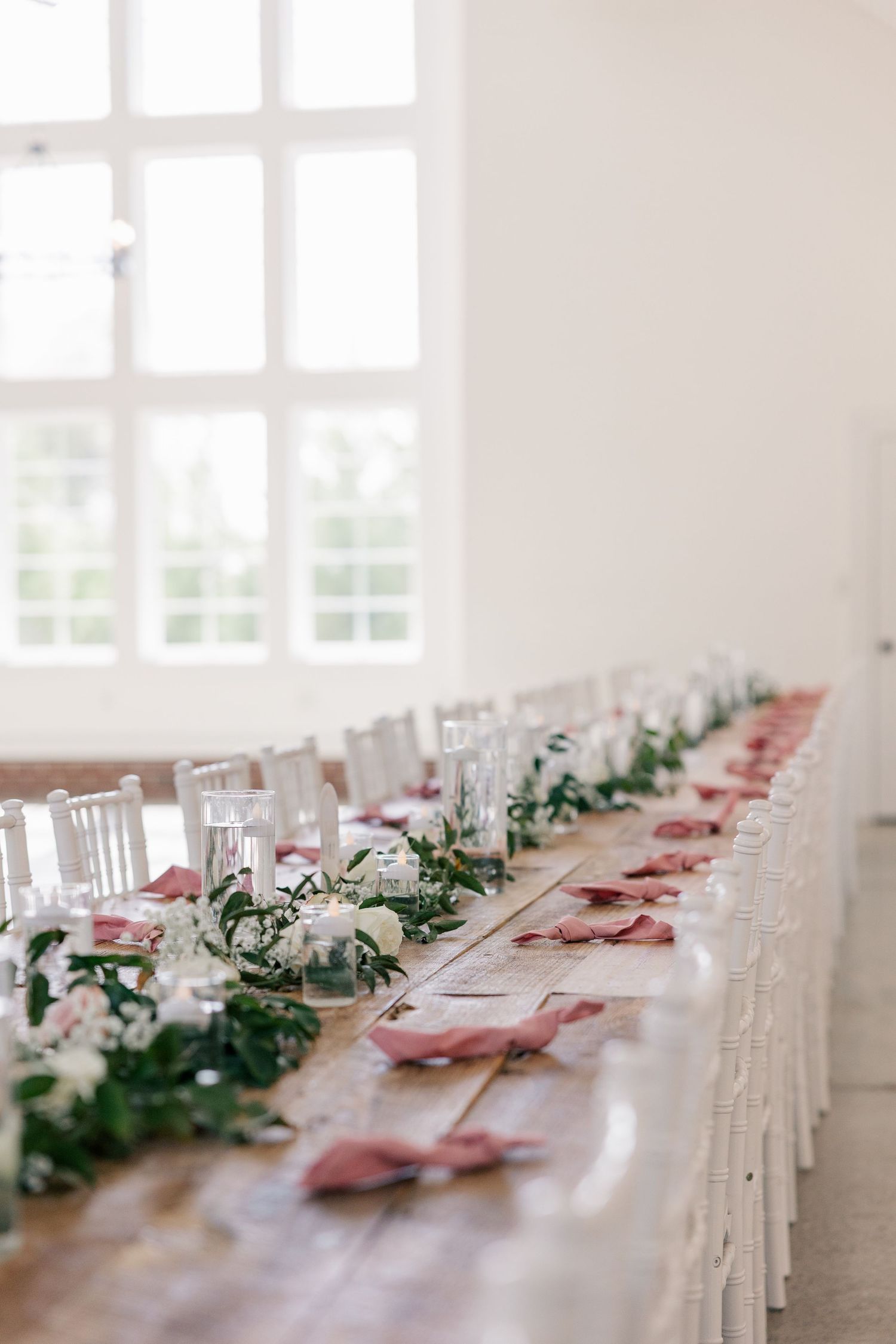 Elegant wedding reception table setting with pink napkins and greenery garland decorations in a bright white room.