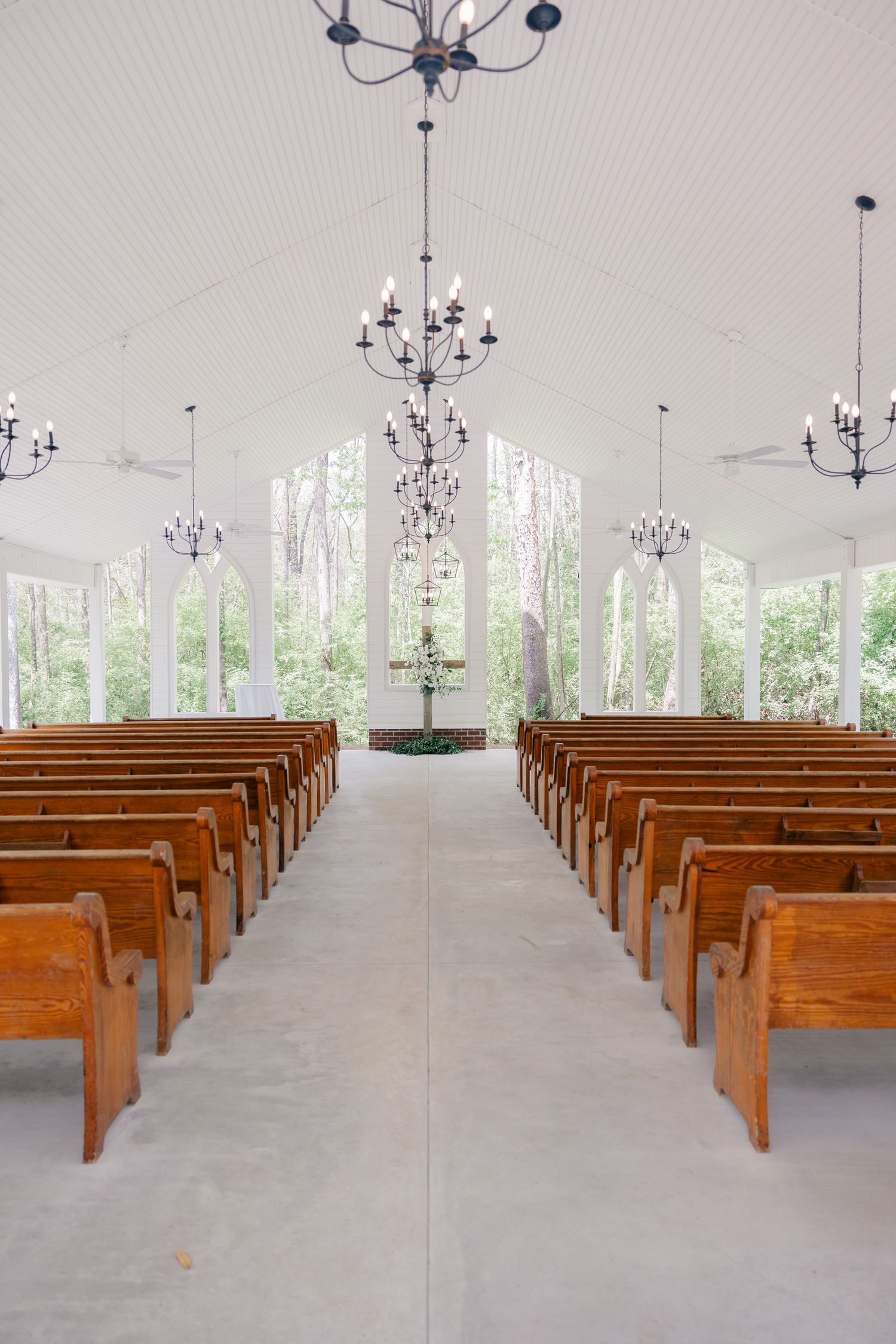 Long rows of wooden pews in a bright chapel with chandeliers and floor-to-ceiling windows.