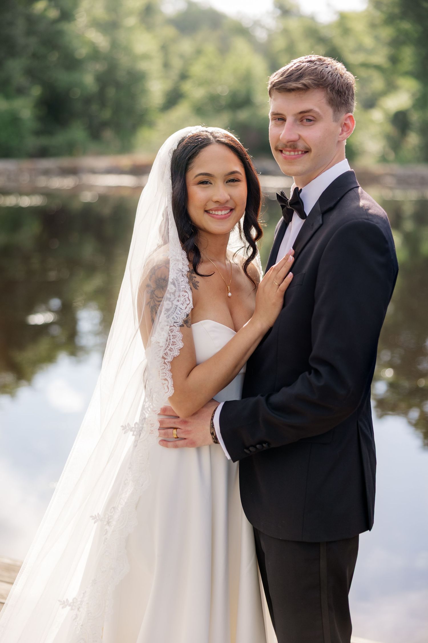 Newlywed couple poses by pond with bride in lace veil and white gown.