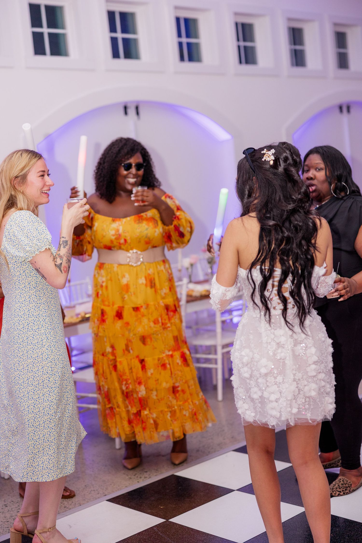 Wedding guests dancing and celebrating in short dresses on a white dance floor with purple accent lighting.