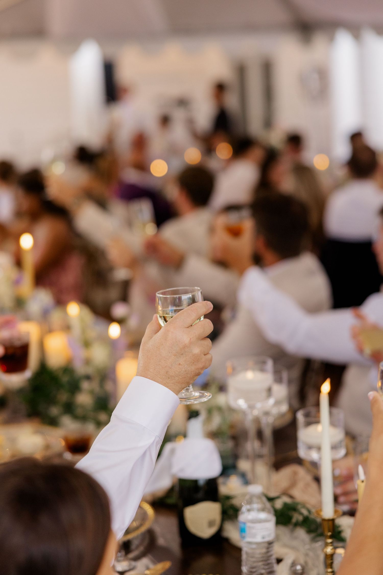 Wedding reception dinner table with glowing candles and raised glasses in a dimly lit banquet hall.