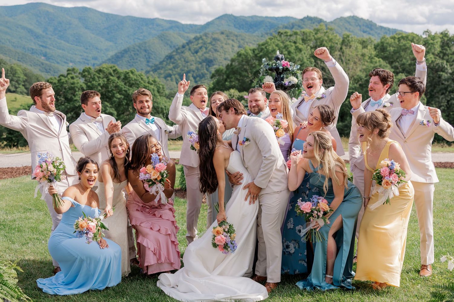 Wedding party celebrates with joyful poses against a backdrop of lush mountain scenery on a sunny summer day.