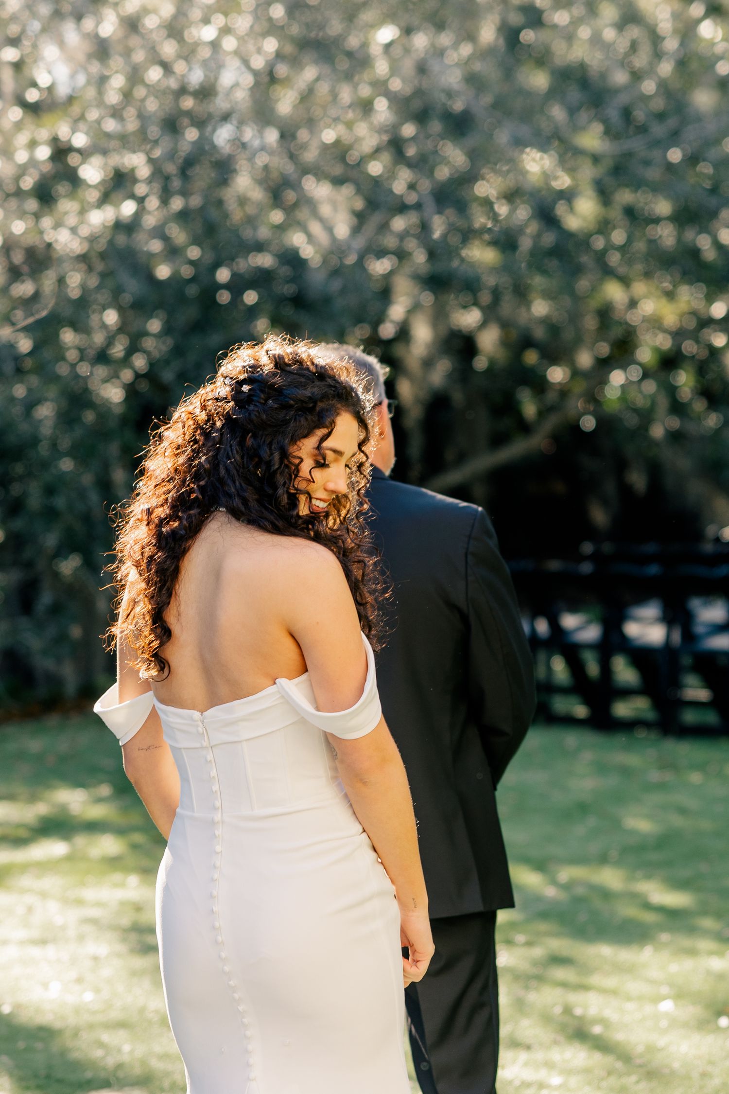 A bride in a fitted white dress stands next to her groom in a garden setting with sunlit bokeh in the background.