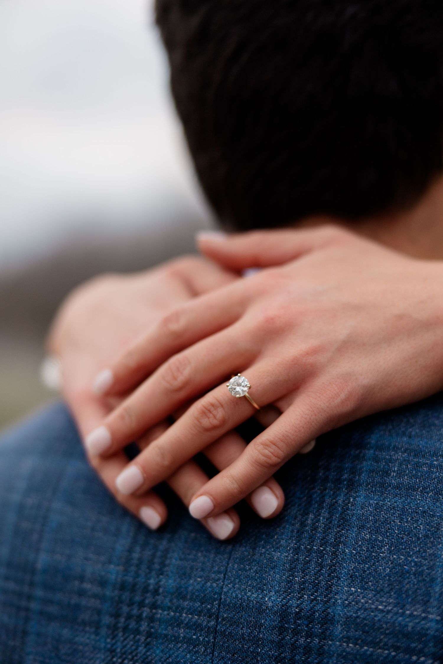 Close-up of hands with diamond engagement ring resting on lap with denim jeans visible in soft lighting.