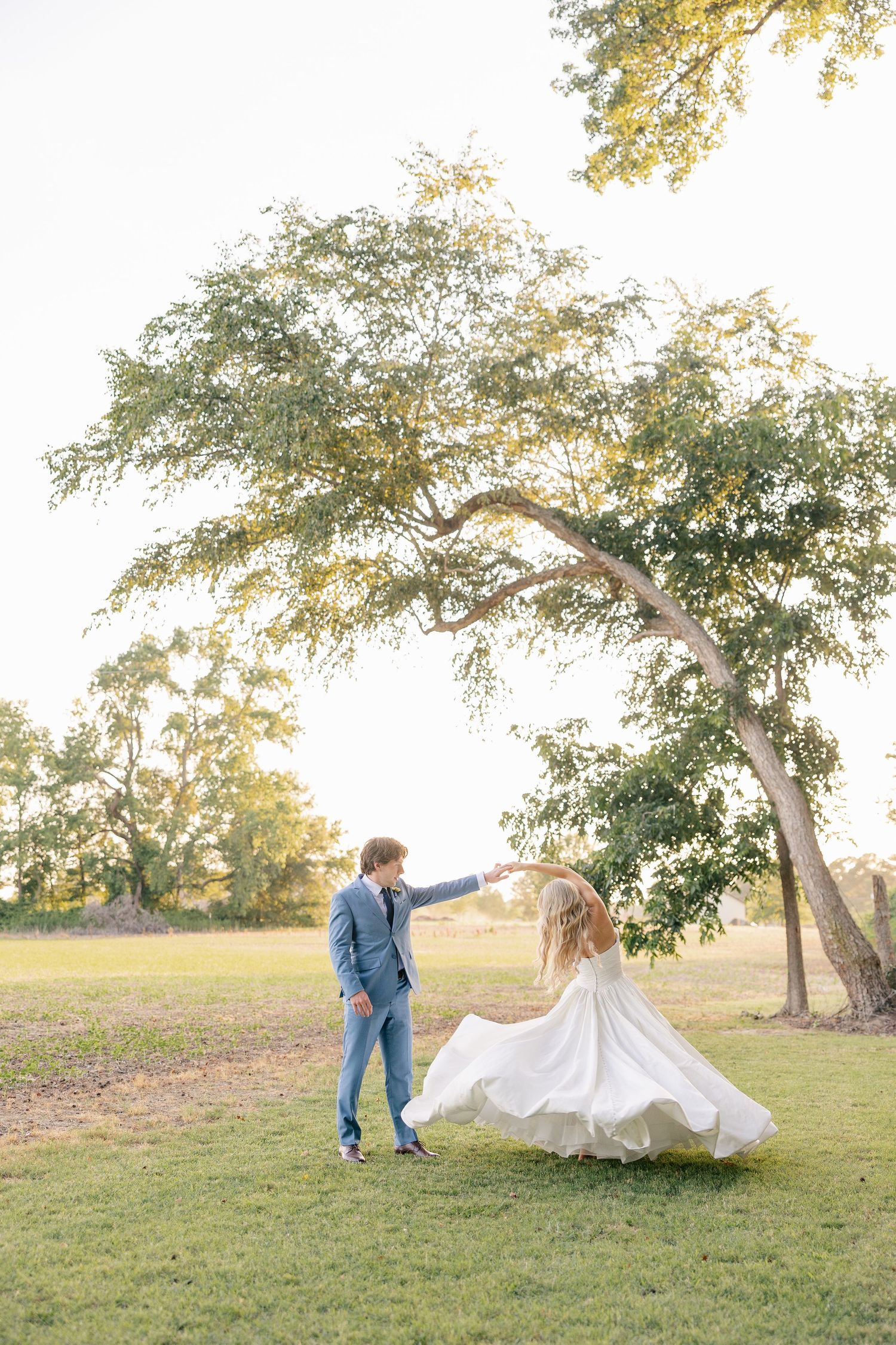 Groom spinning bride in flowing white dress under a large tree at sunset in a green meadow.