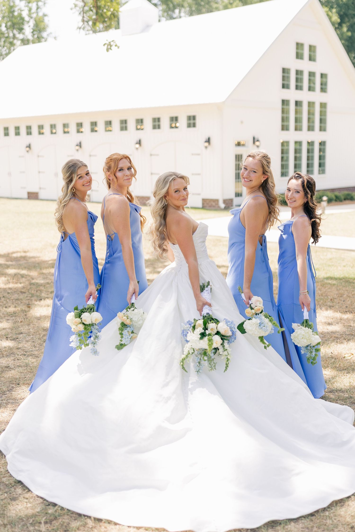 Bride in white gown poses with four bridesmaids in periwinkle blue dresses holding white bouquets outdoors.