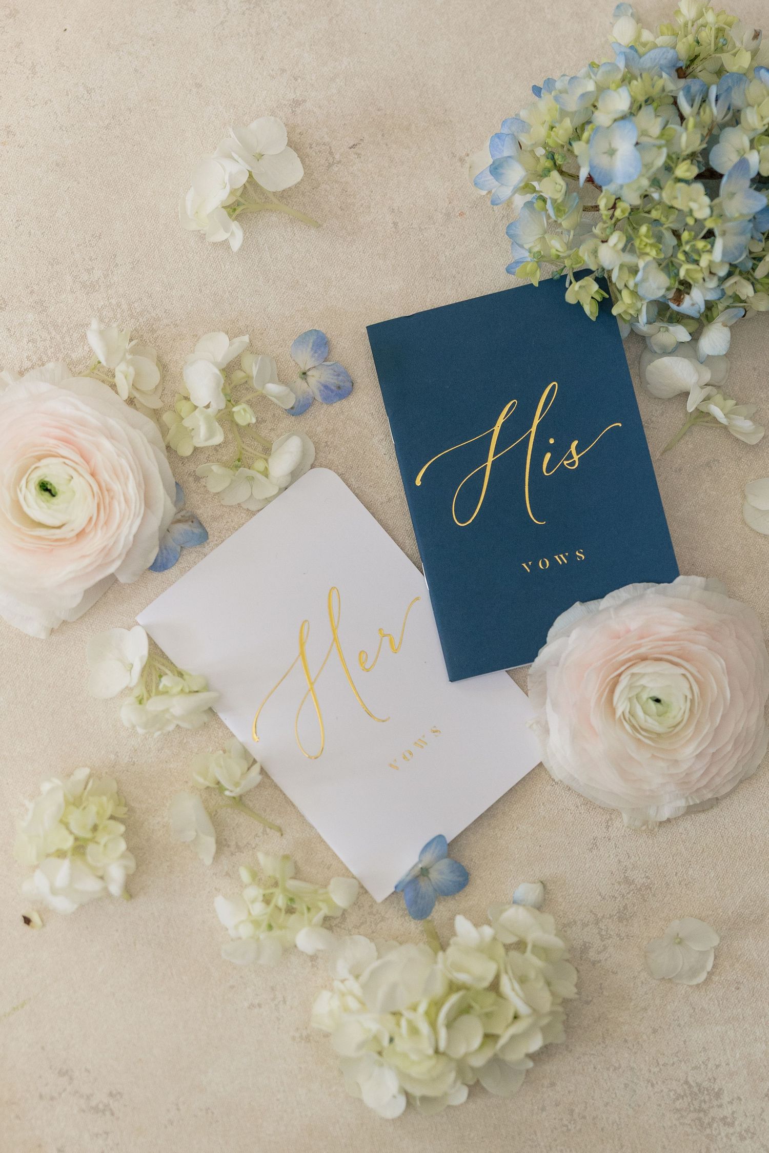 His and Her vow books in navy and white with gold script surrounded by flowers.