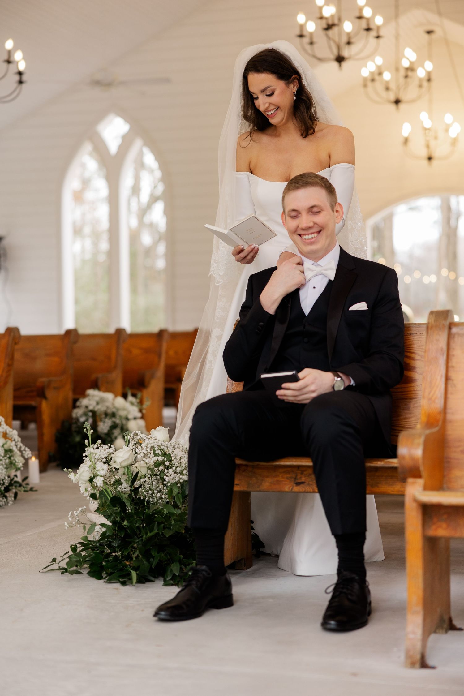 Bride in white gown reads vows while groom in black tuxedo sits smiling in a decorated white chapel.