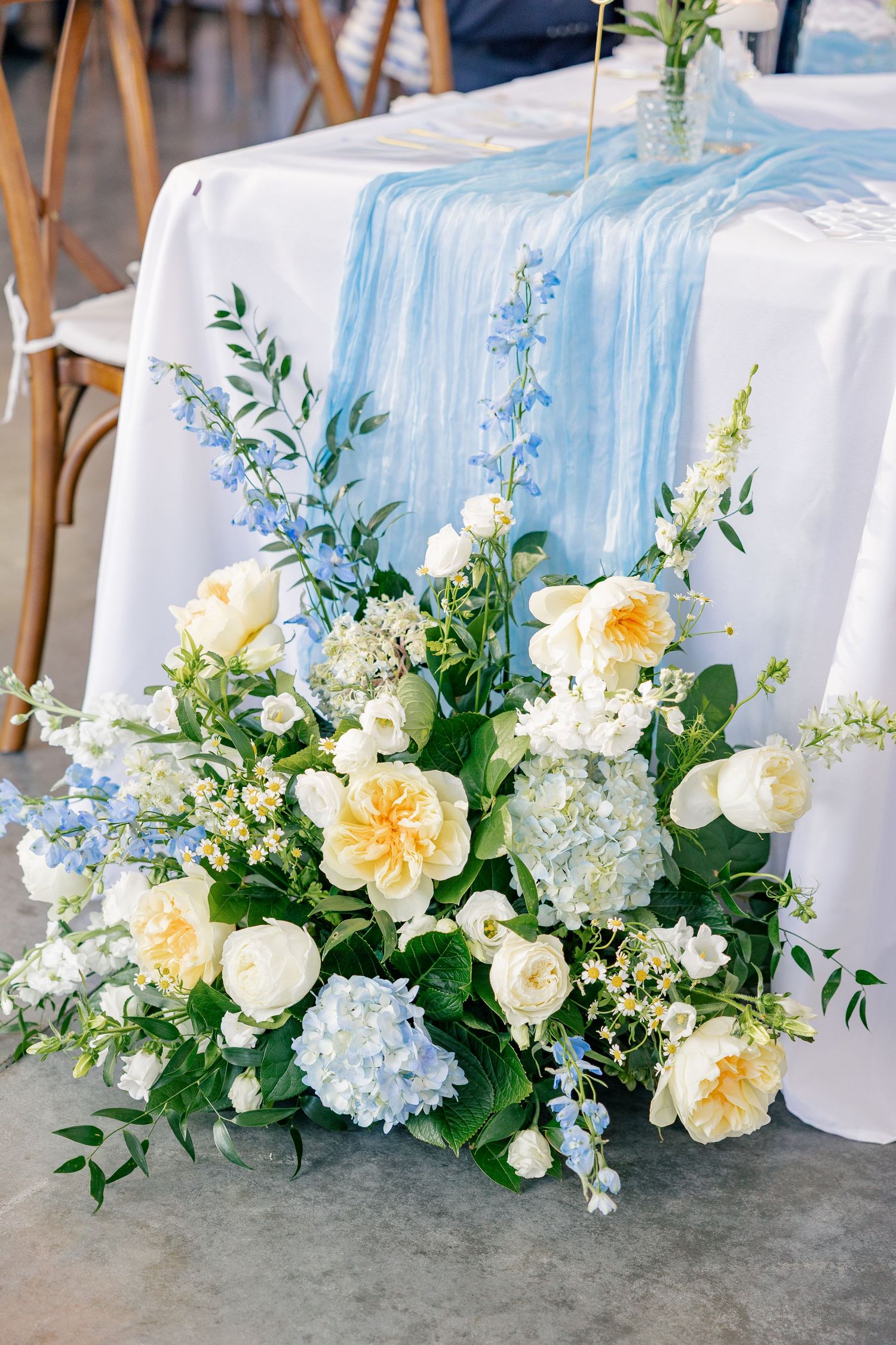 Elegant wedding table arrangement with yellow roses, white blooms, blue hydrangeas and light blue table runner.