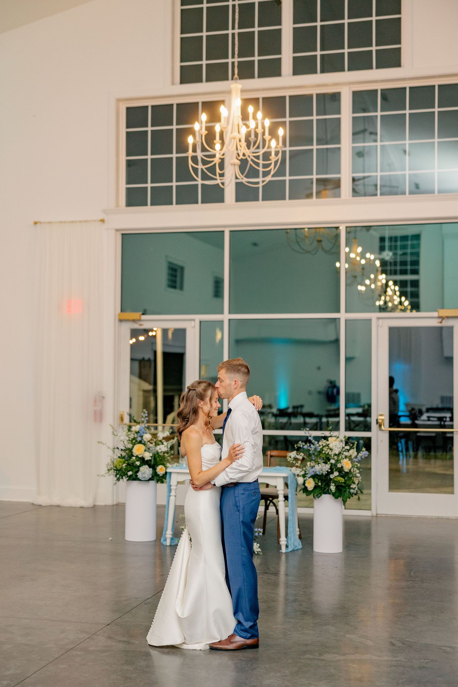 Bride and groom share first dance under chandelier in elegant white wedding venue.